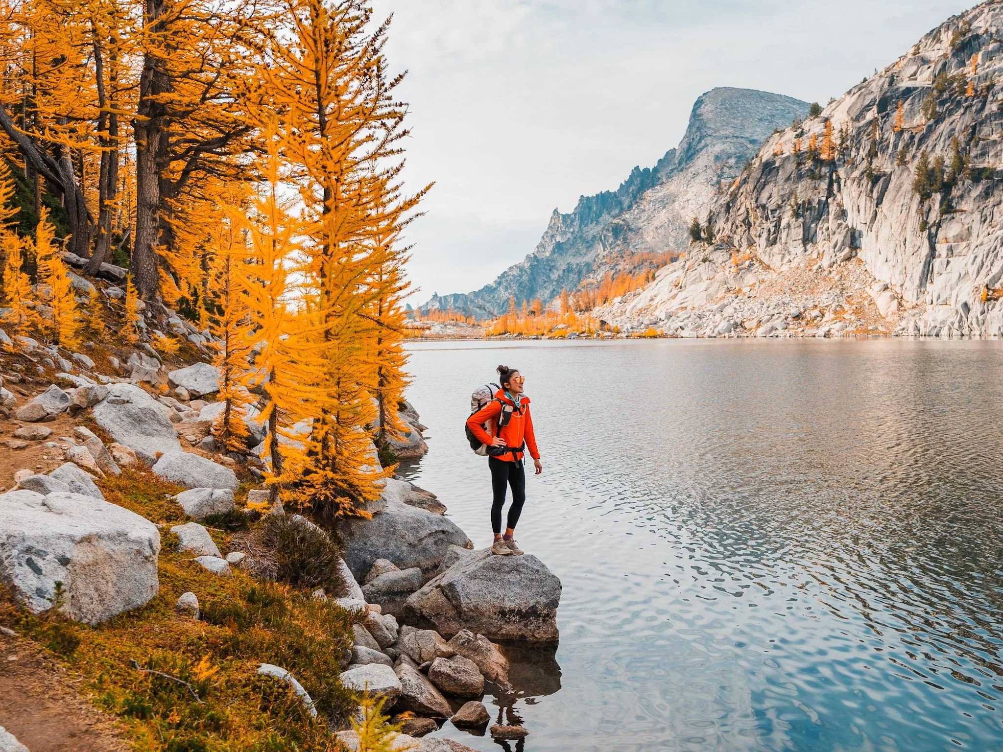 a woman standing in front of a lake in the enchantments, wearing a red jacket, with golden larches and mountain views