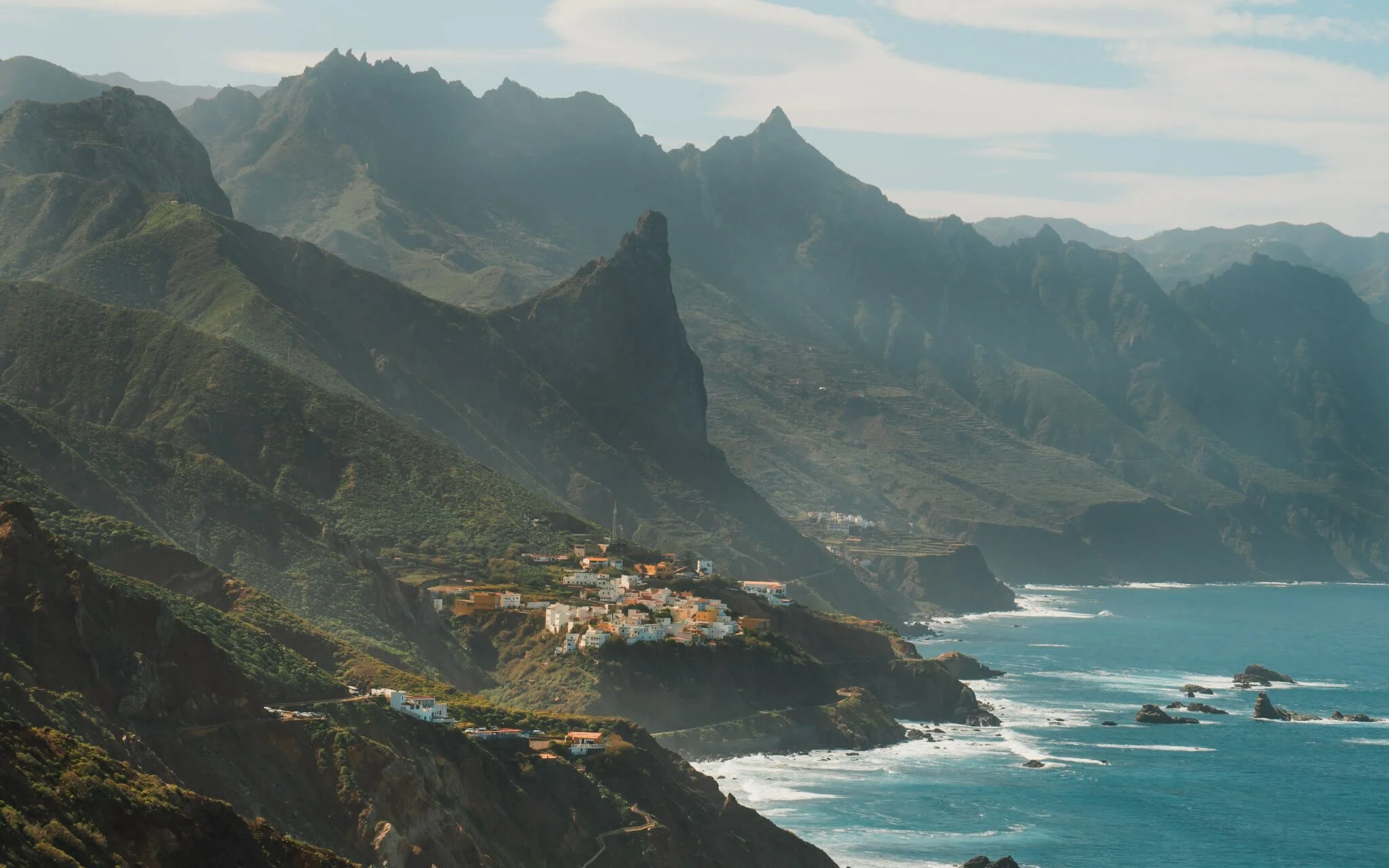 a small town sitting on the edge of a cliff, overlooking the ocean, with large green hills and cliffs in the background
