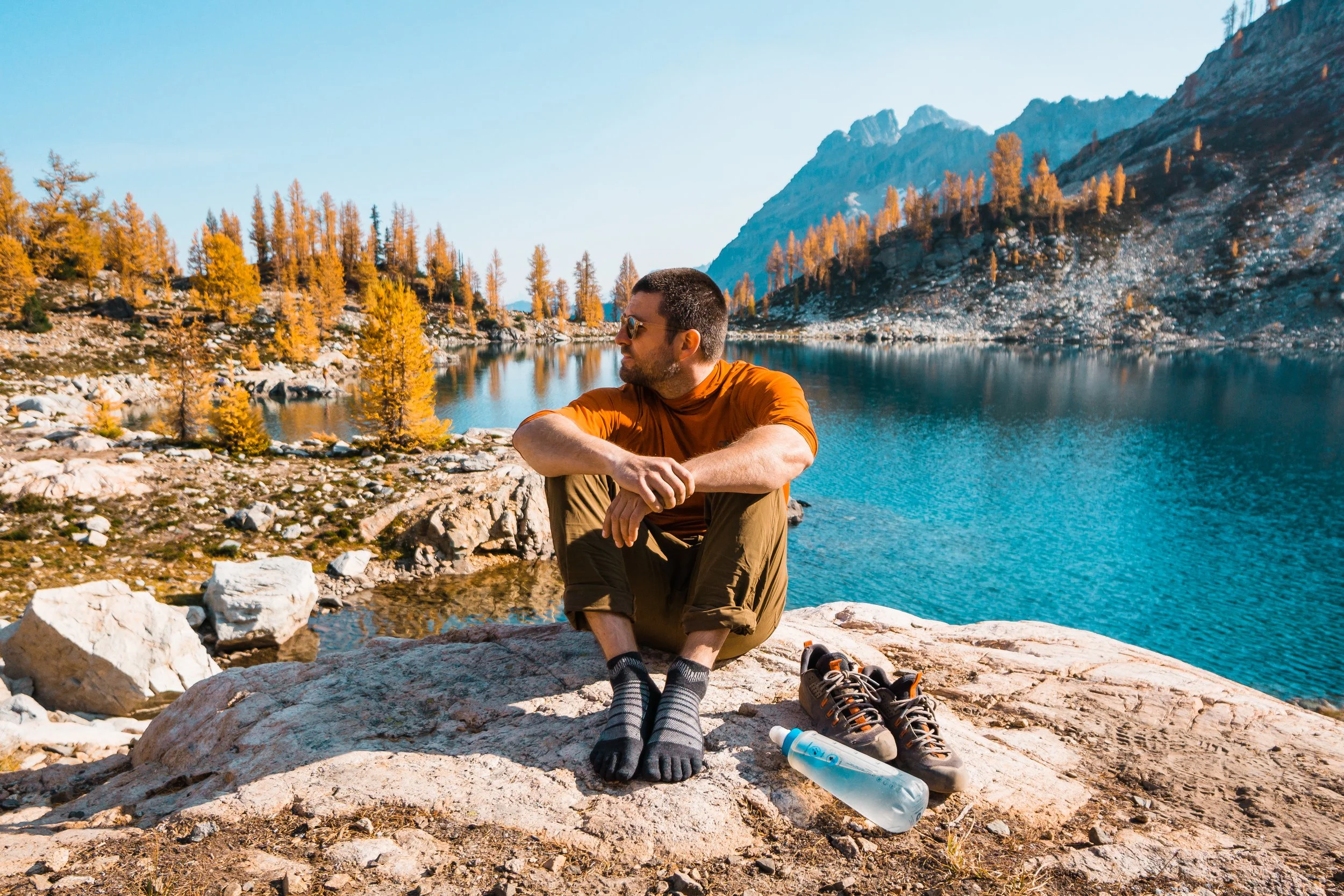 Man sitting on a rock near a lake with mountains in the background, wearing sunglasses, an orange shirt, and khaki pants, with hiking shoes and a water bottle nearby.