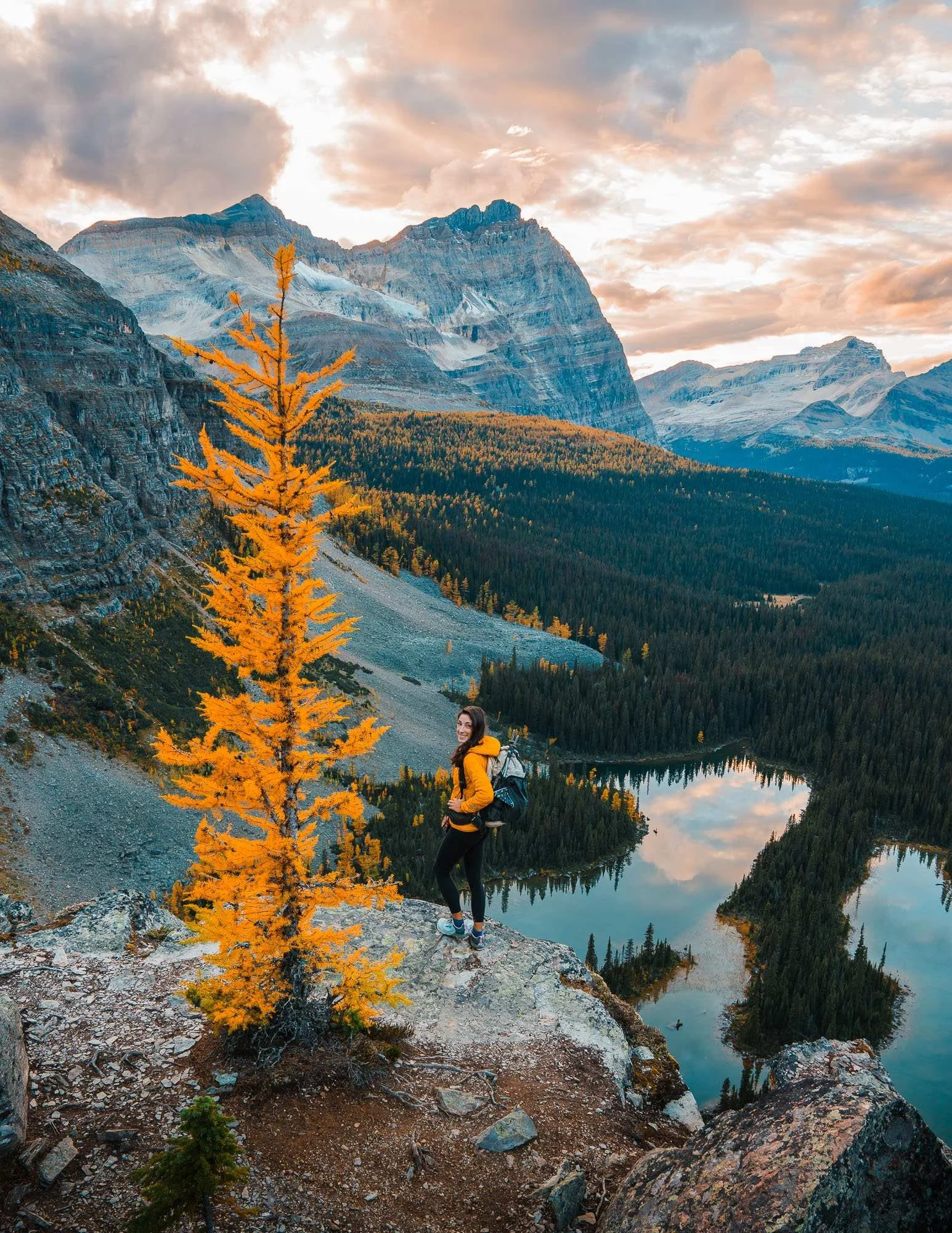 A woman with a backpack standing on a rocky ledge overlooking a lake surrounded by trees and mountains, with a partly cloudy sky, during autumn.