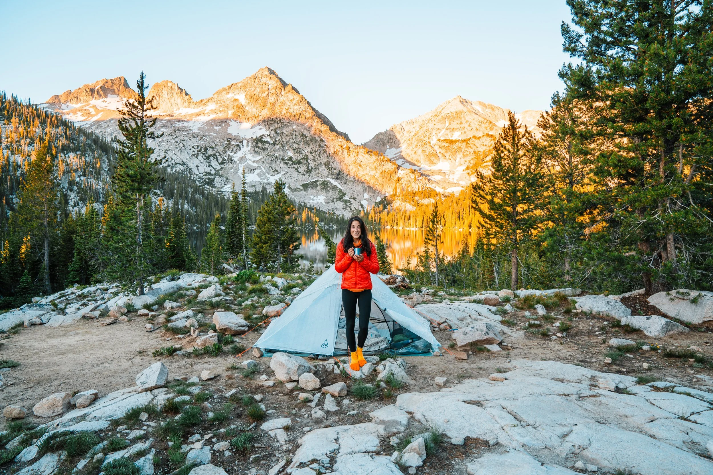 a woman standing in front of a white tent, holding a coffee, while backpacking in the idaho sawtooth mountains