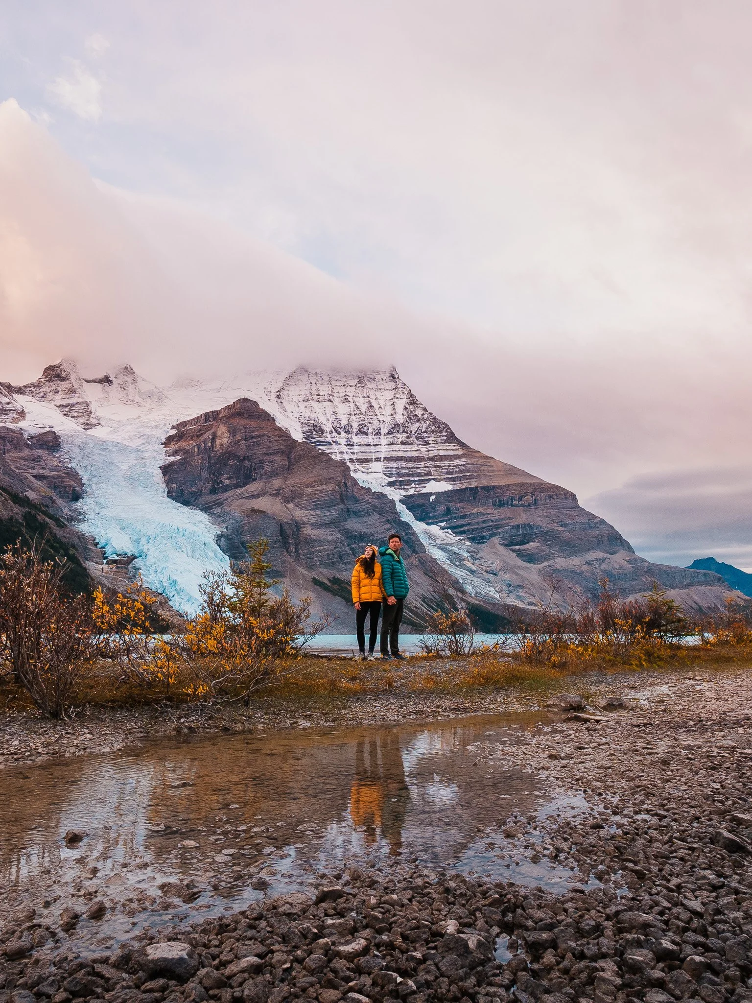 A man and woman stand together outdoors near a body of water with mountains and glaciers in the background, wearing warm jackets.
