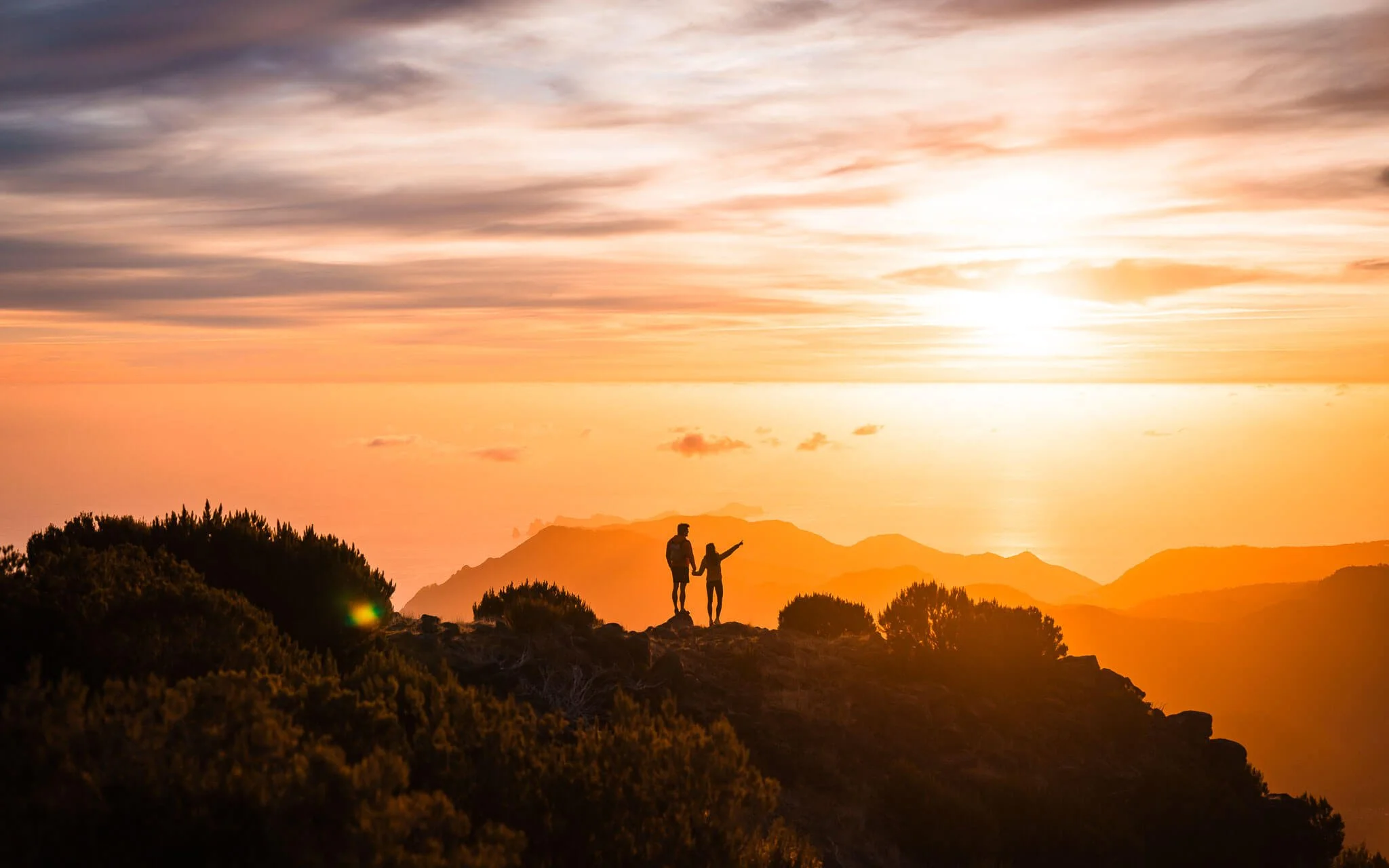 silhouettes of a couple looking out at the ocean while on a hike, with a bright orange sunrise happening