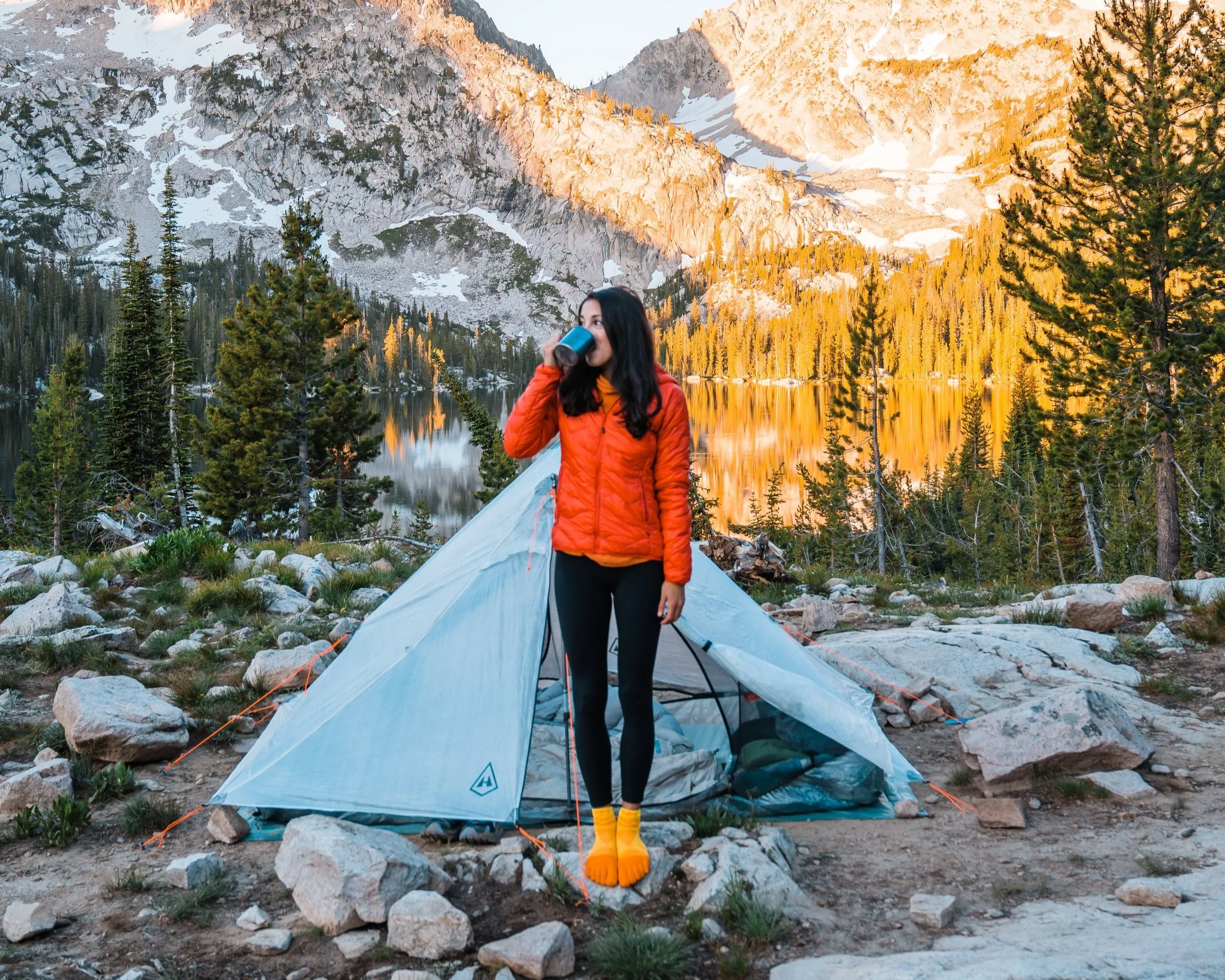 A woman in an orange jacket and yellow socks standing outside a tent by a lake in a mountain landscape, drinking from a blue mug.