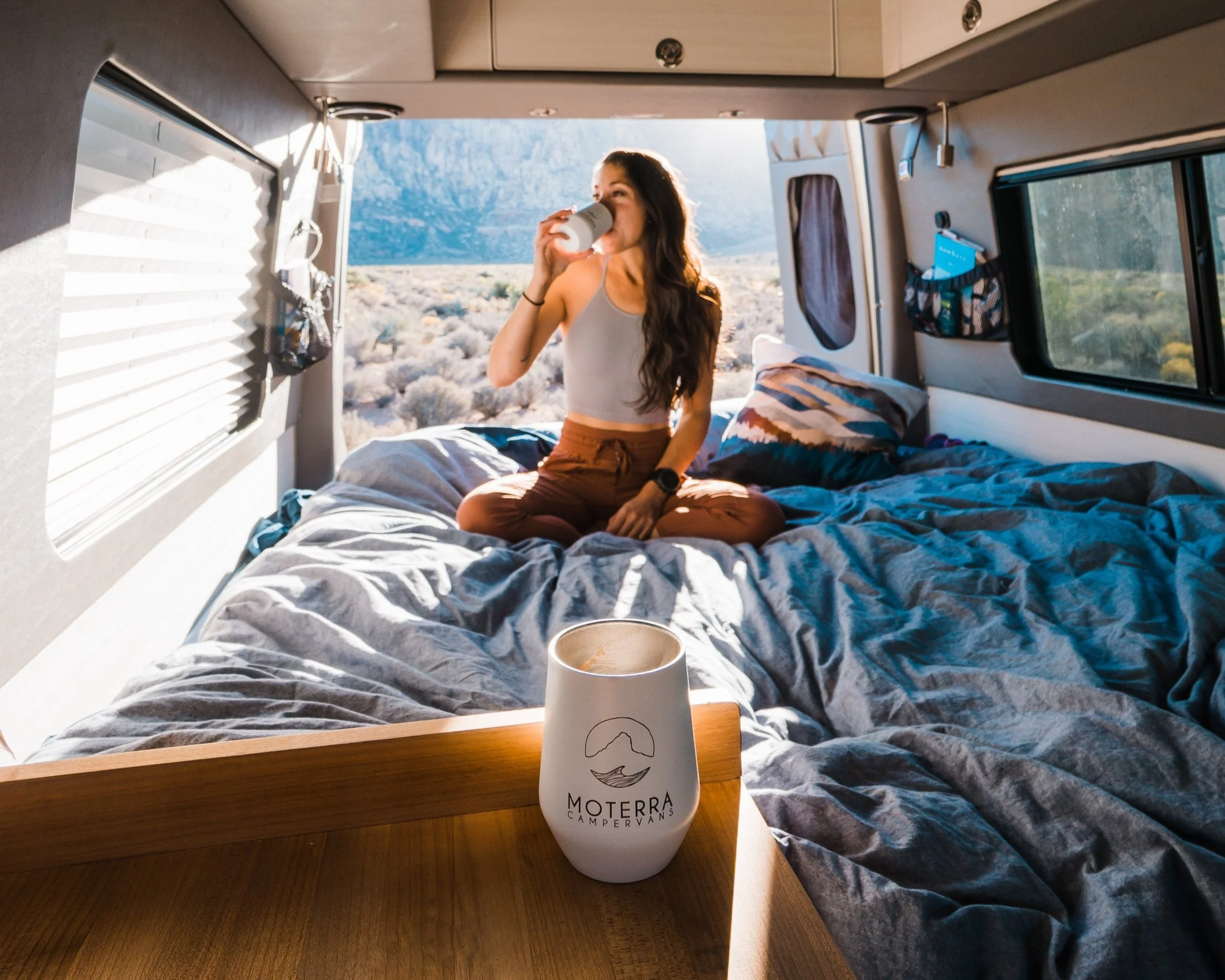 A woman sitting cross-legged inside an RV, drinking from a mug, with a view of a desert landscape and mountains outside the window.