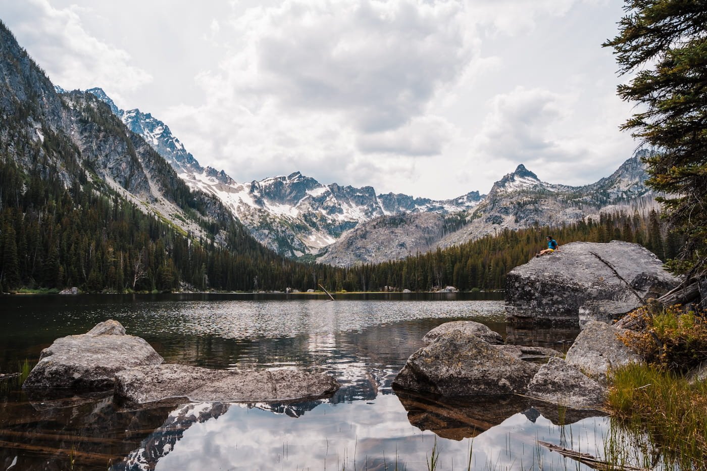 a tiny hiker sitting on a big boulder at the edge of lake stuart in the enchantments