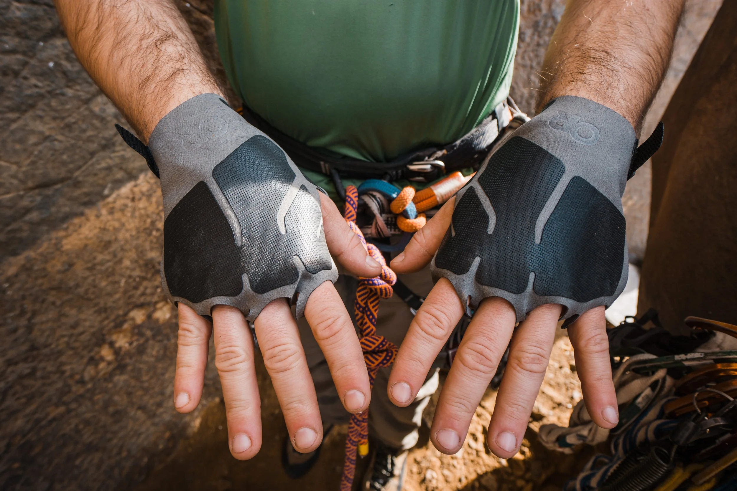 Close-up of a person wearing climbing gloves with padded palms, showing the person's hands and part of their green shirt and climbing harness, with a rocky outdoor background.