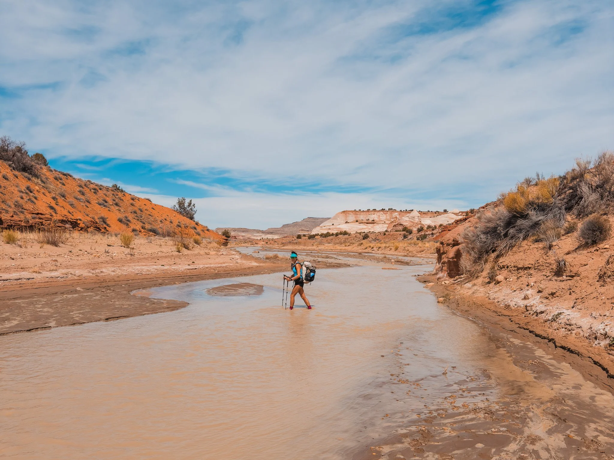 a woman wearing a white hyperlite backpacking pack and shorts, crossing a muddy creek with trekking poles in the desert