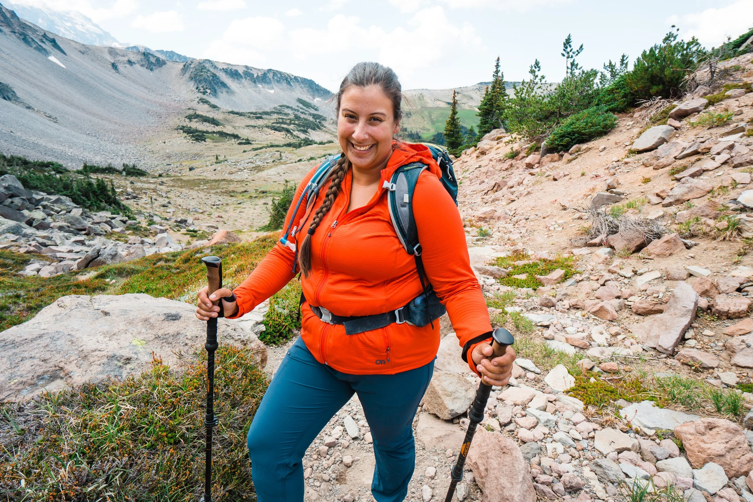 A woman hiking on a rocky trail in a mountainous area with a backpack, smiling, holding hiking poles with mountains in the background.