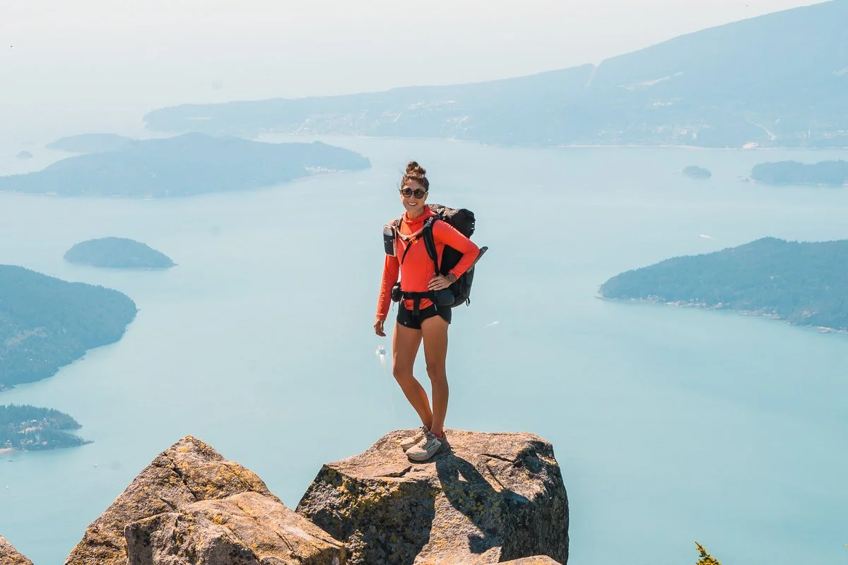 a woman standing in front of the howe sound in british columbia, wearing a bright pink sun hoddie and black backpack on the howe sound crest trail