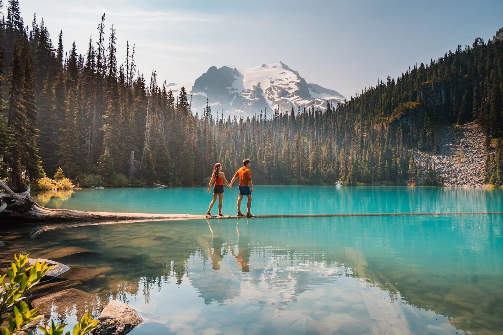 Couple walking across a floating log in the middle of a bright blue alpine lake (Middle Joffre Lake) with mountains in the background