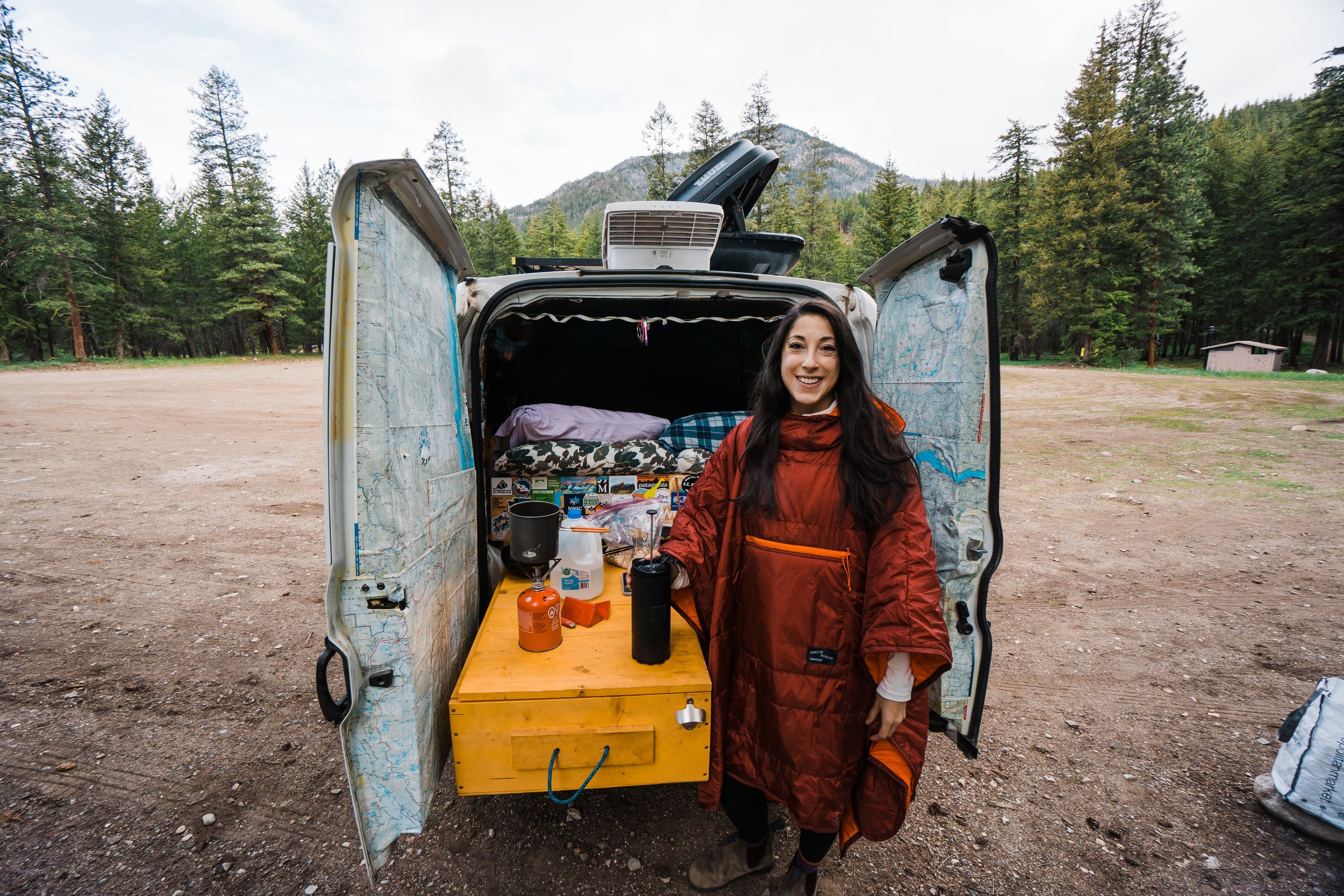 Woman in a red camping coat standing outside an open van with a camping setup, surrounded by trees in a forested area.