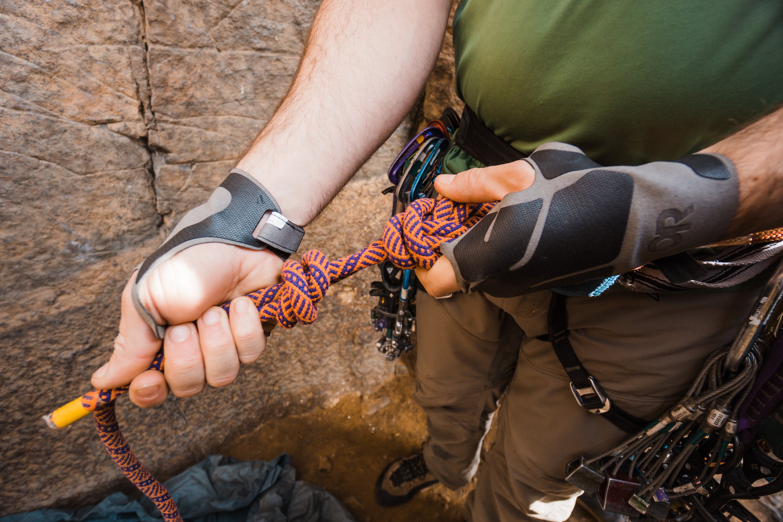 Close-up of two people shaking hands, one wearing climbing gloves and the other wearing a wrist brace, with climbing gear and carabiners attached to their harnesses, in front of a rock wall.