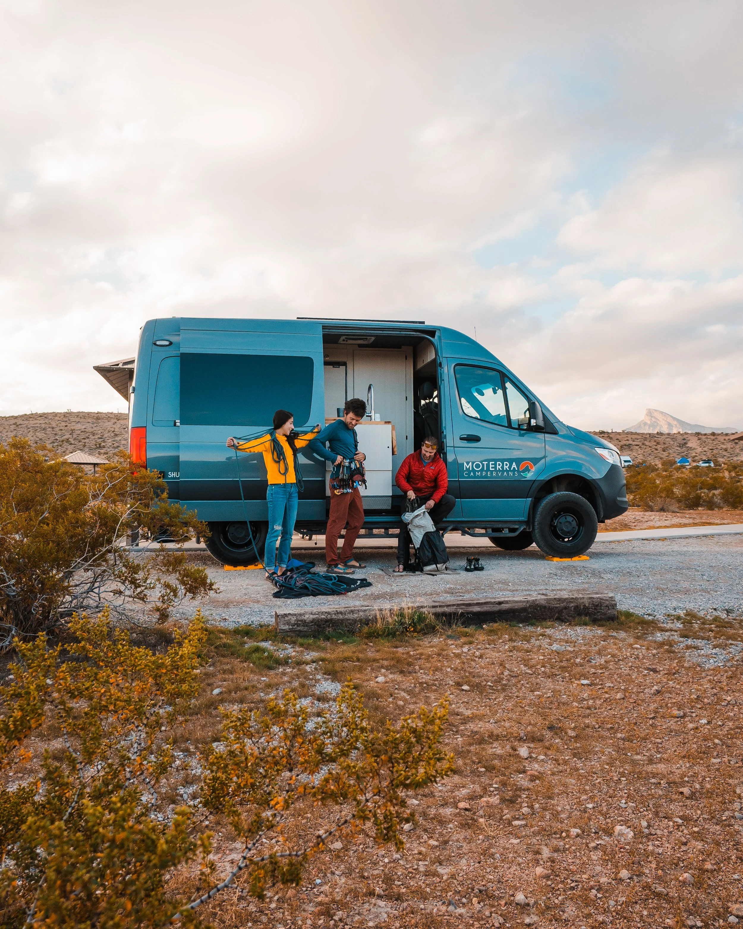 Three people preparing gear next to a blue camper van in a desert landscape with sparse bushes and mountains in the background.