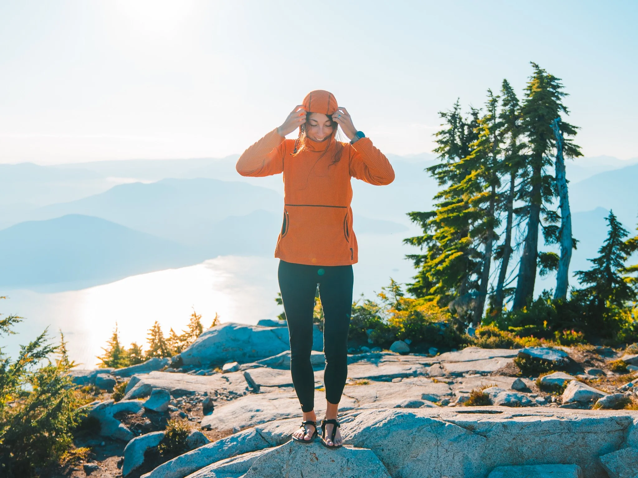 A woman in an orange grid fleece hoodie, with the hood up, standing in front of some costal views