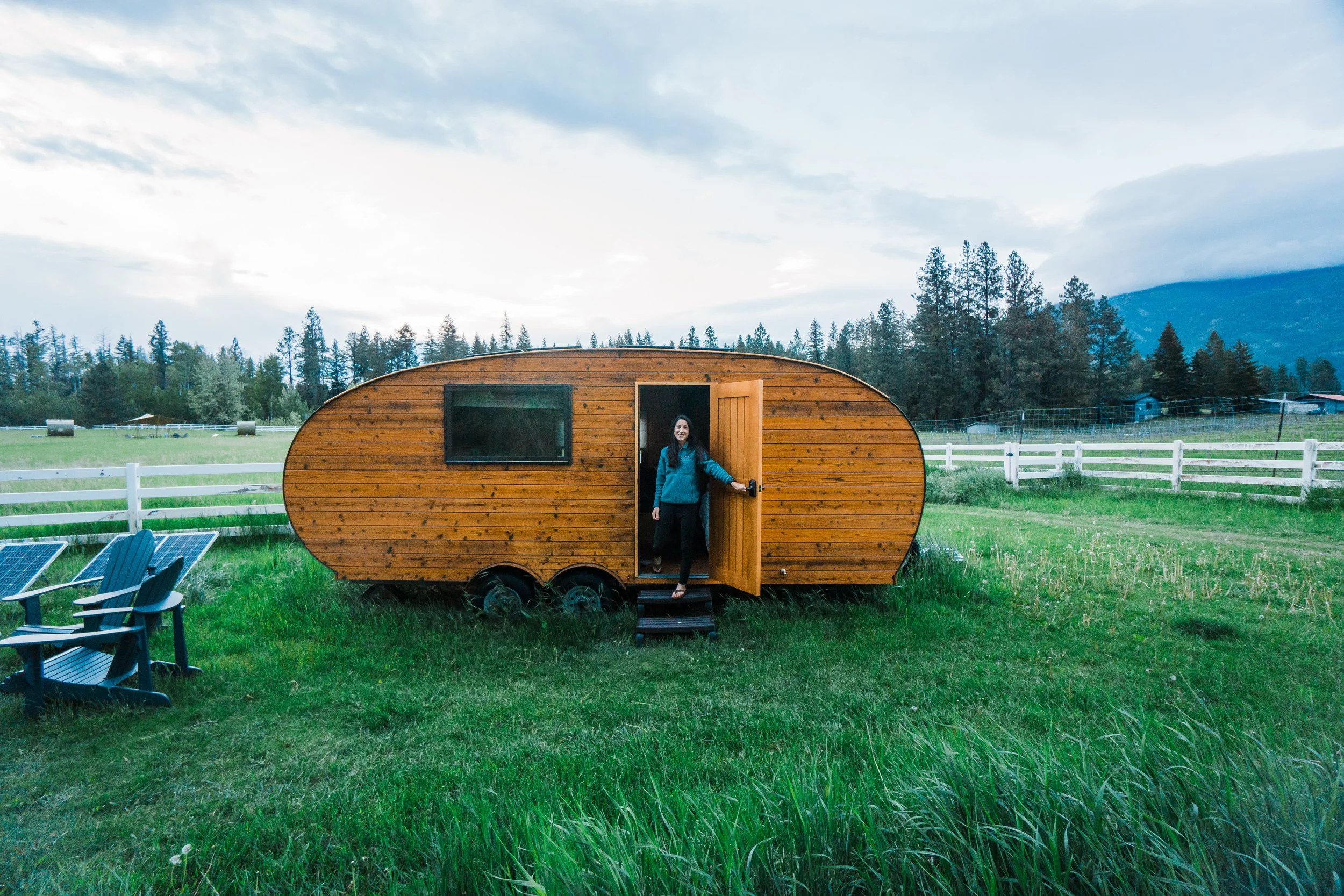 Woman standing at the entrance of a small wooden travel trailer in a grassy field with a white fence, trees, mountains, and a cloudy sky in the background.