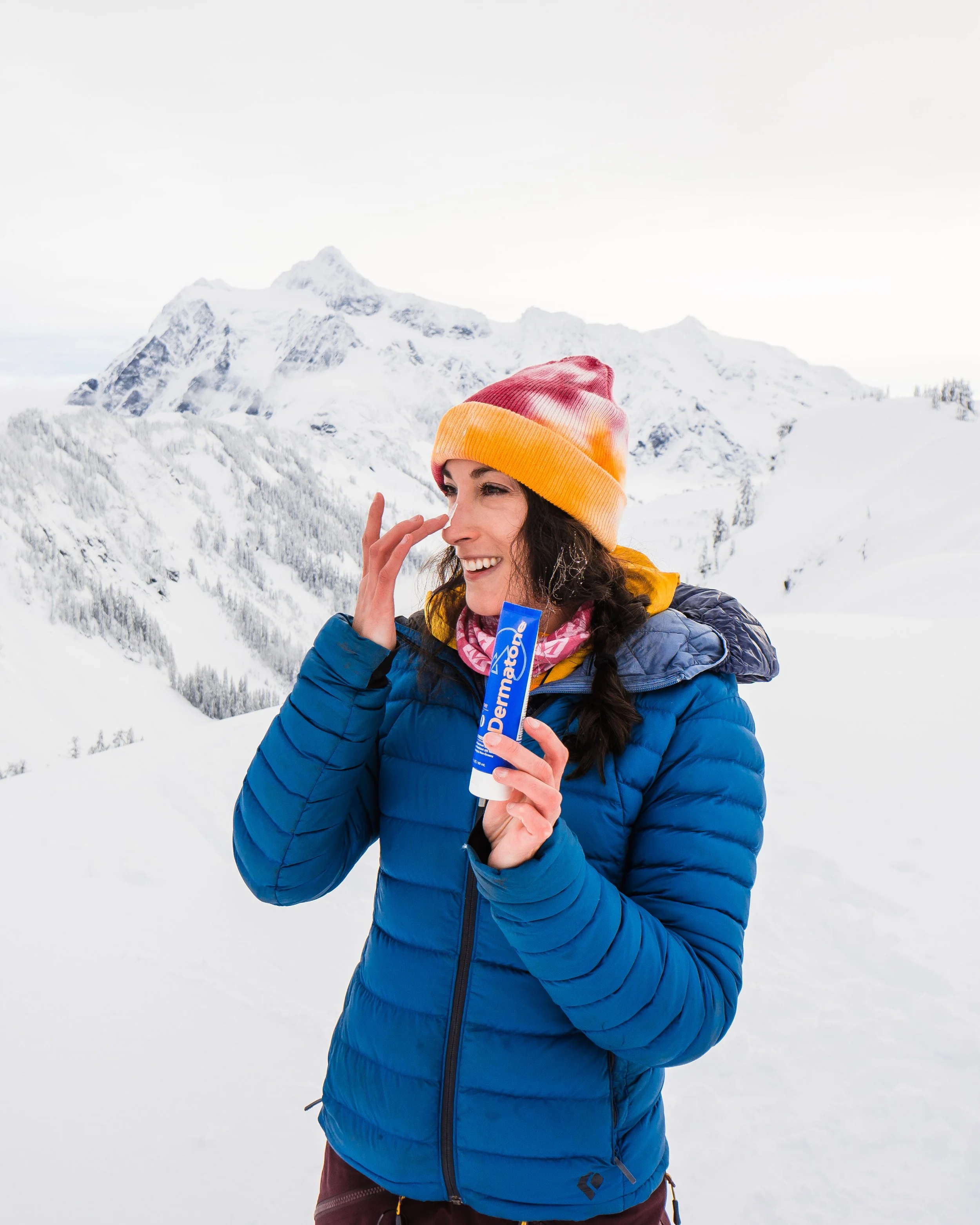 A woman in a colorful beanie and blue jacket holding sunscreen in a snowy mountain landscape.
