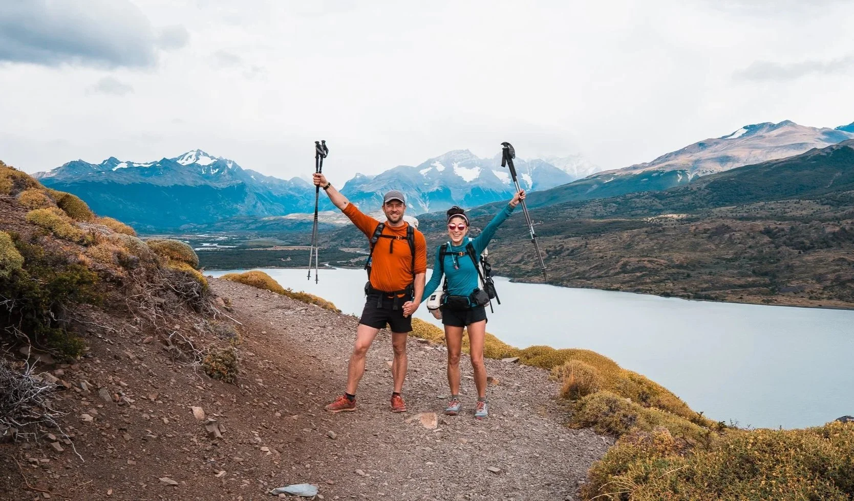 Two hikers, a man and a woman, standing on a mountain trail with a lake and mountains in the background. They are smiling, holding hiking poles, and wearing backpacks.