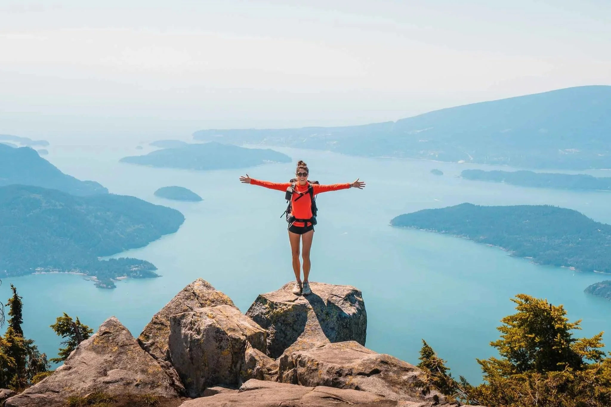 a woman standing on a rock in front of the howe sound with arms outstretched while backpacking