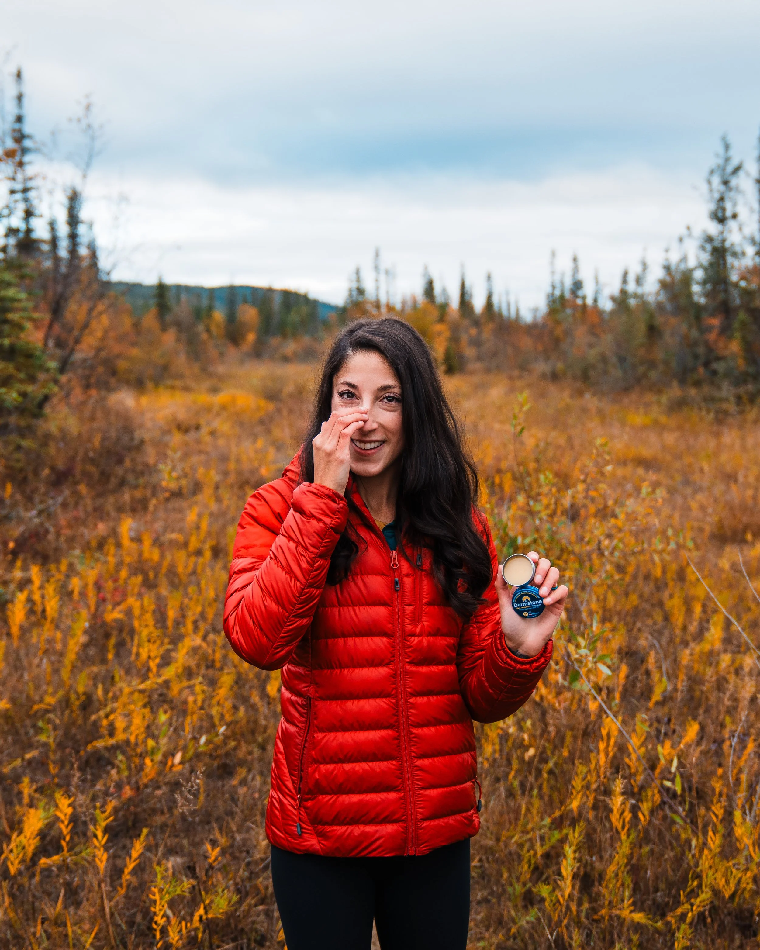 A woman in a red jacket holding lip balm and smiling in an autumn landscape with orange and yellow foliage.
