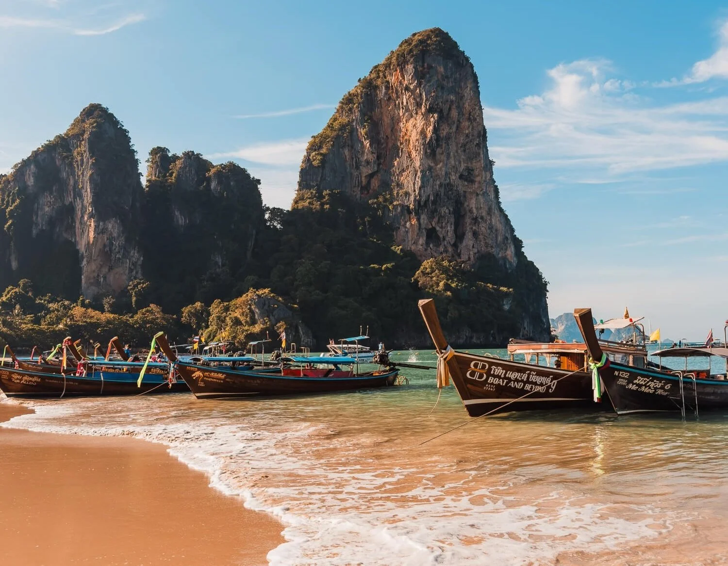 View of Railay beach in krabi, thailand with orange sand, blue water, long tail boats, and a massive rock face in the background.
