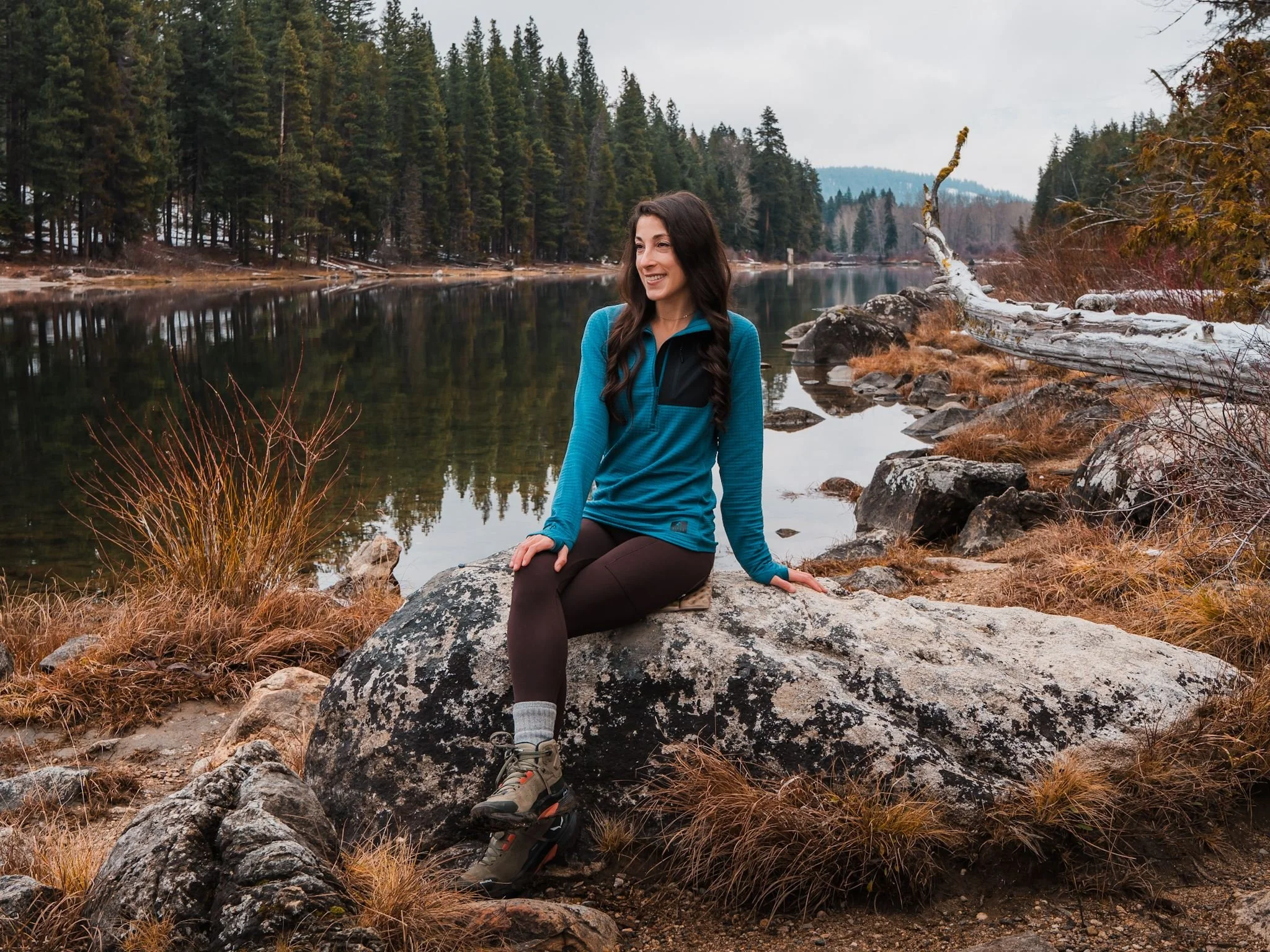 A woman sitting on a rock in front of a lake with pine trees in the background. She is wearing a blue fleece and dark leggings