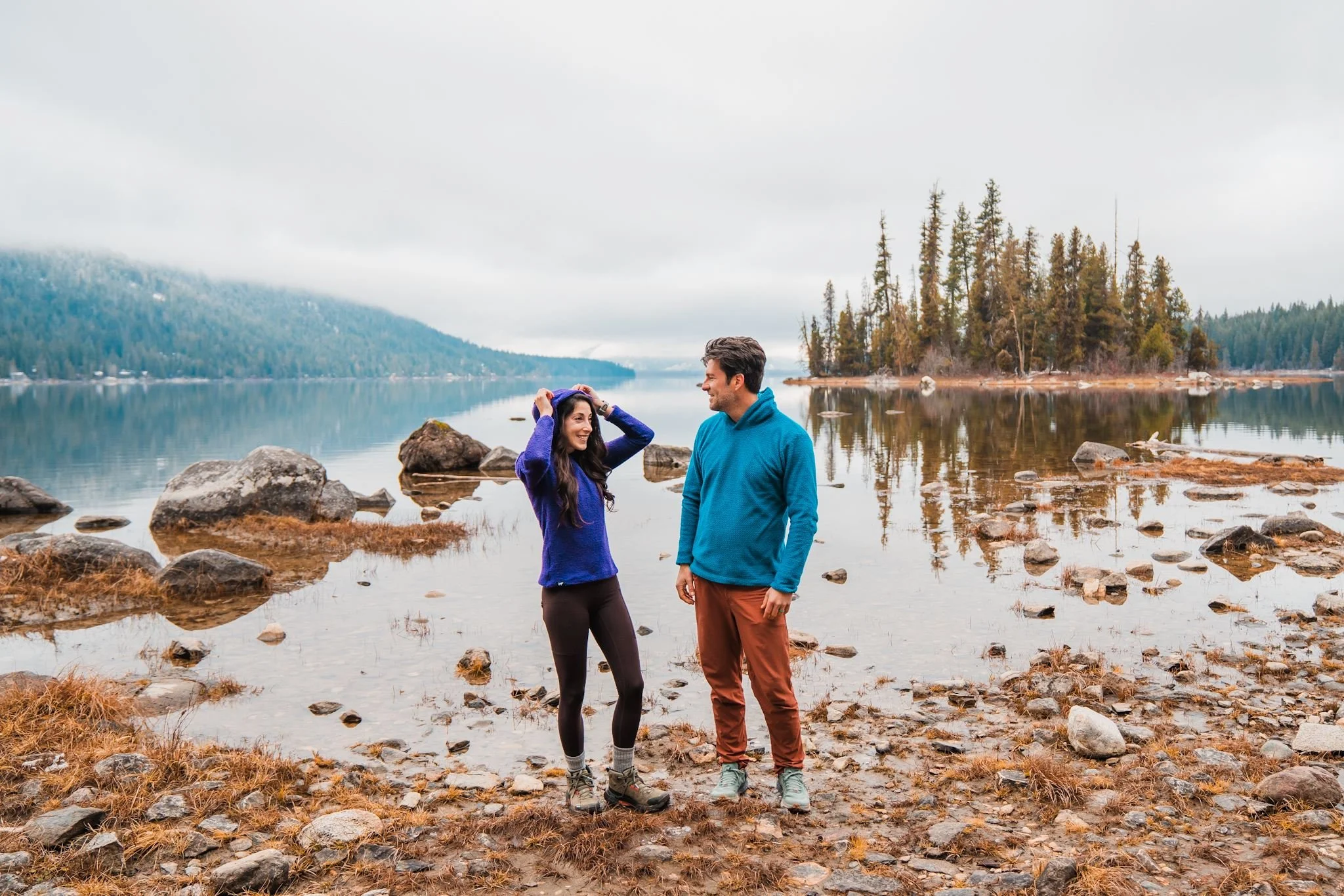 a couple standing in front of a lake with forested island in the background, wearing alpha direct fleece hoodies