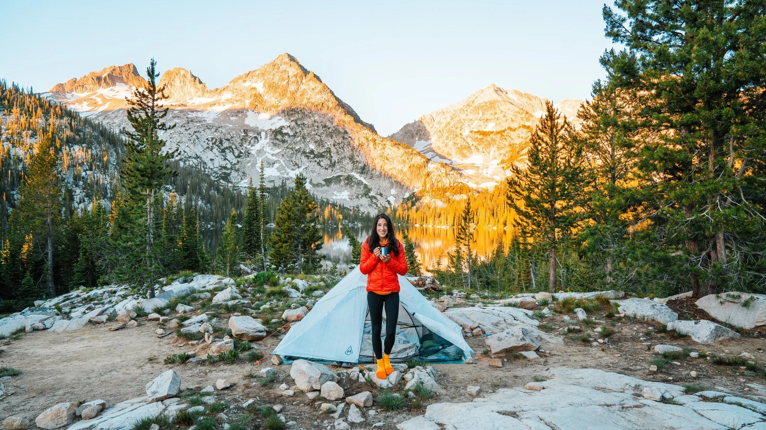 girl holding a camping mug in front of a tent and campsite in the sawtooth mountains
