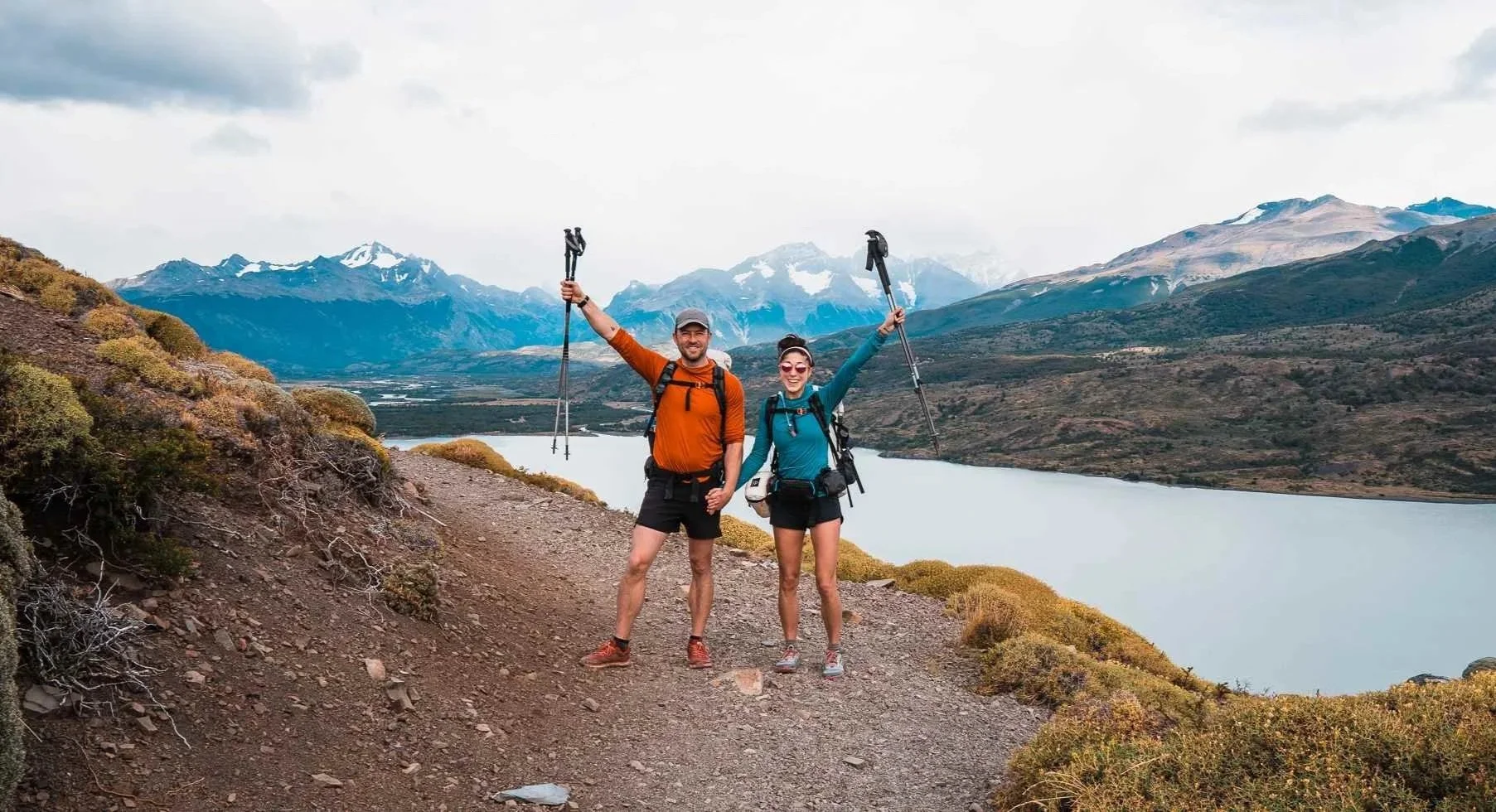 a couple of hikers standing on a trail with arms holding their trekking poles up, wearing bright sun hoodies in patagonia