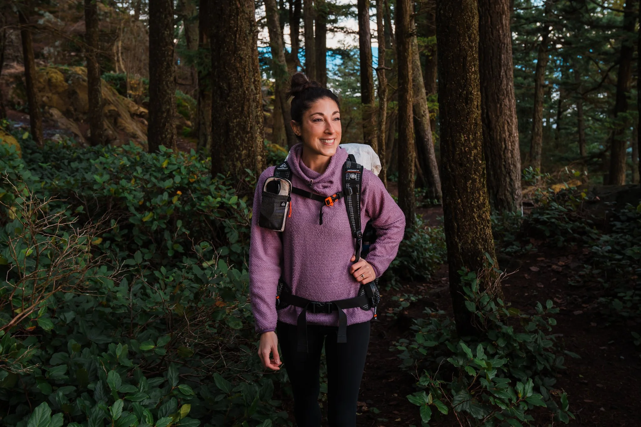 a woman hiking through a forest, wearing a light violet colored fleece hoodie and a backpack
