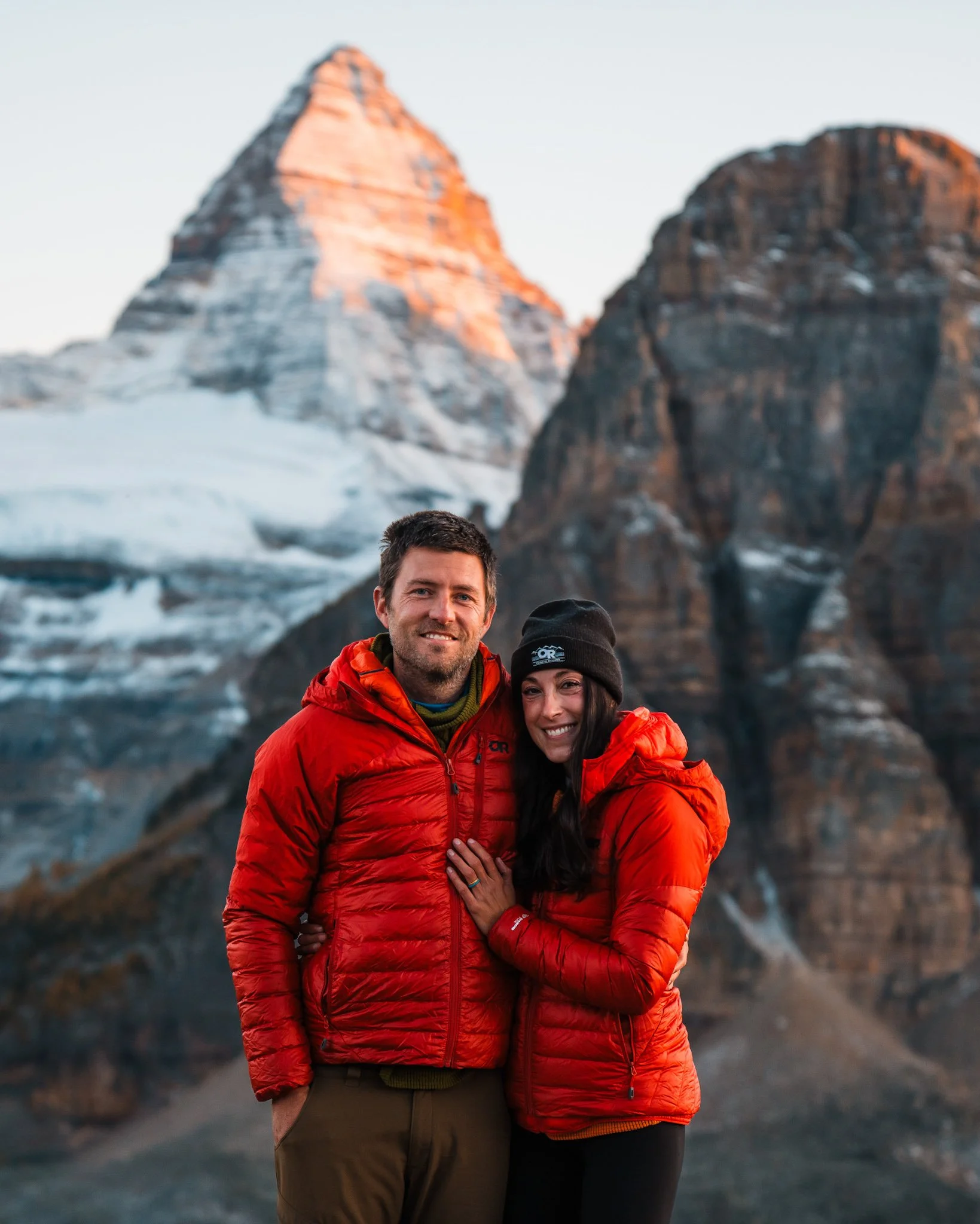 a couple in matching red down jackets, standing in front of big mountains with alpenglow on them at sunset