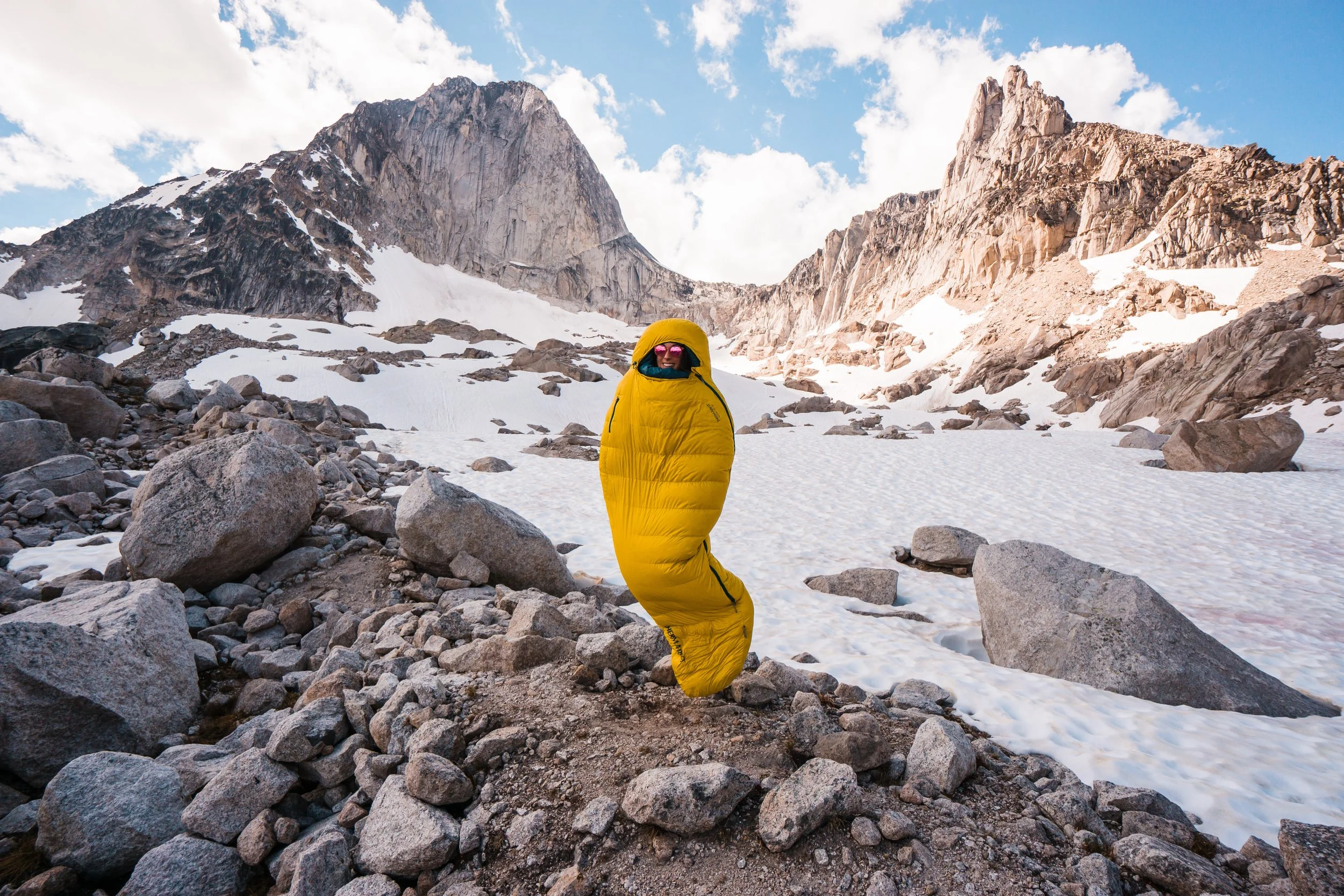 A person in a yellow sleeping bag suit standing on rocky terrain in a snowy mountain landscape with peaks and clouds in the background.