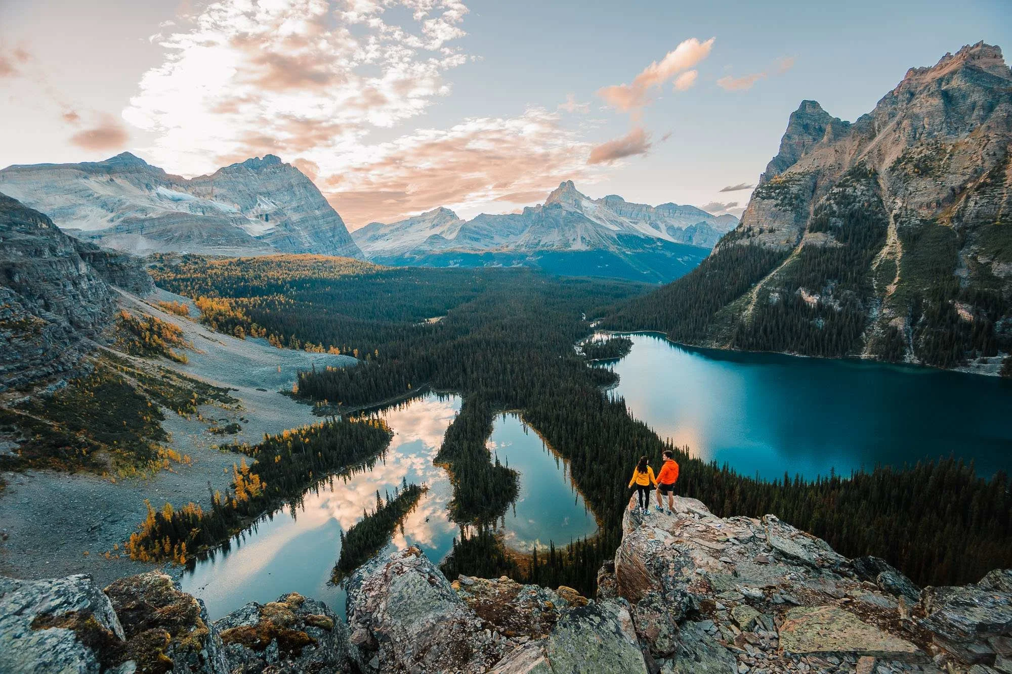 a couple in bright jackets, standing on a cliff overlooking alpine lakes and mountains while the sun is setting over Lake O'Hara on the alpine circuit hike