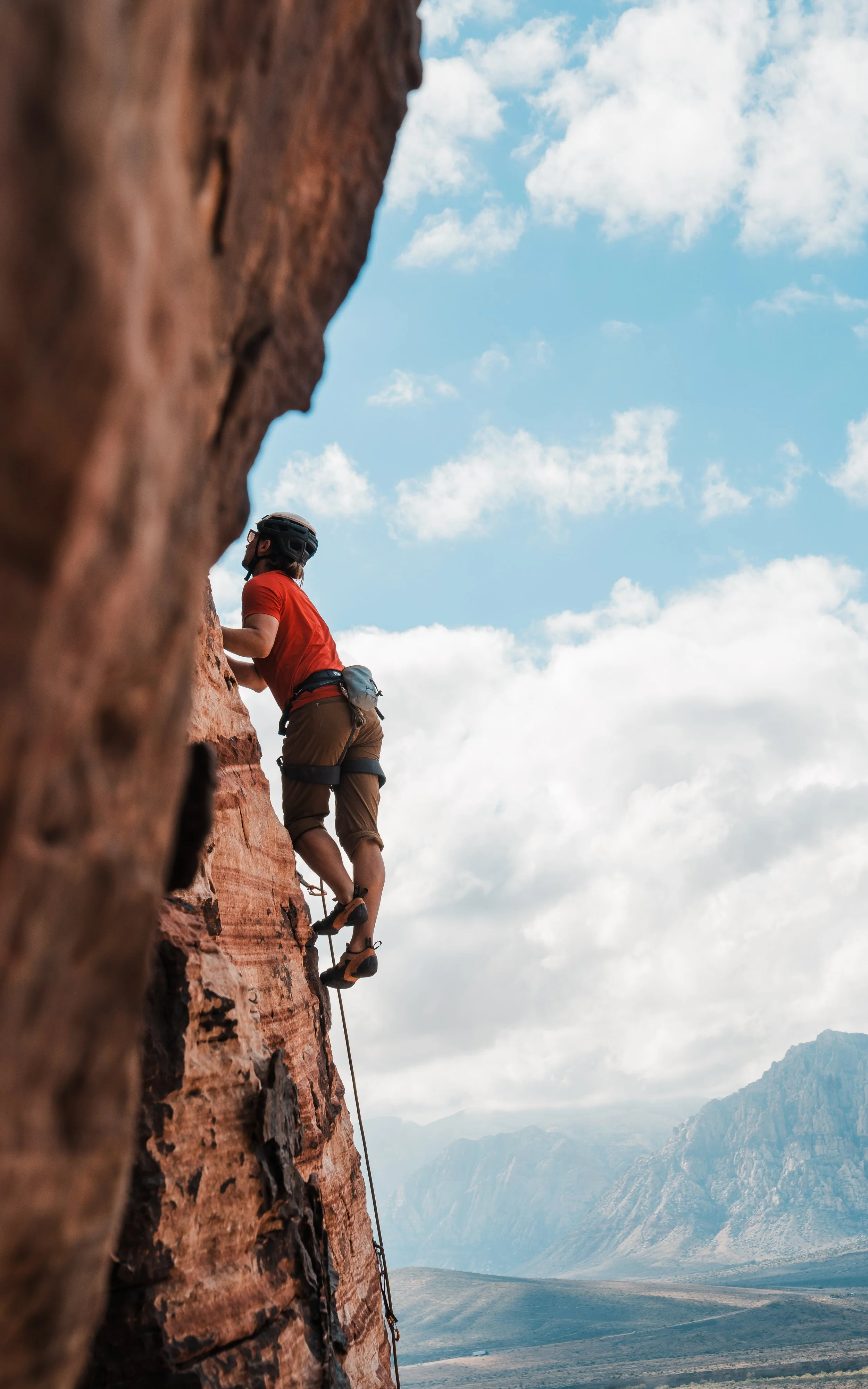 A person rock climbing outdoors on a red rock face under a partly cloudy sky with mountains in the background.