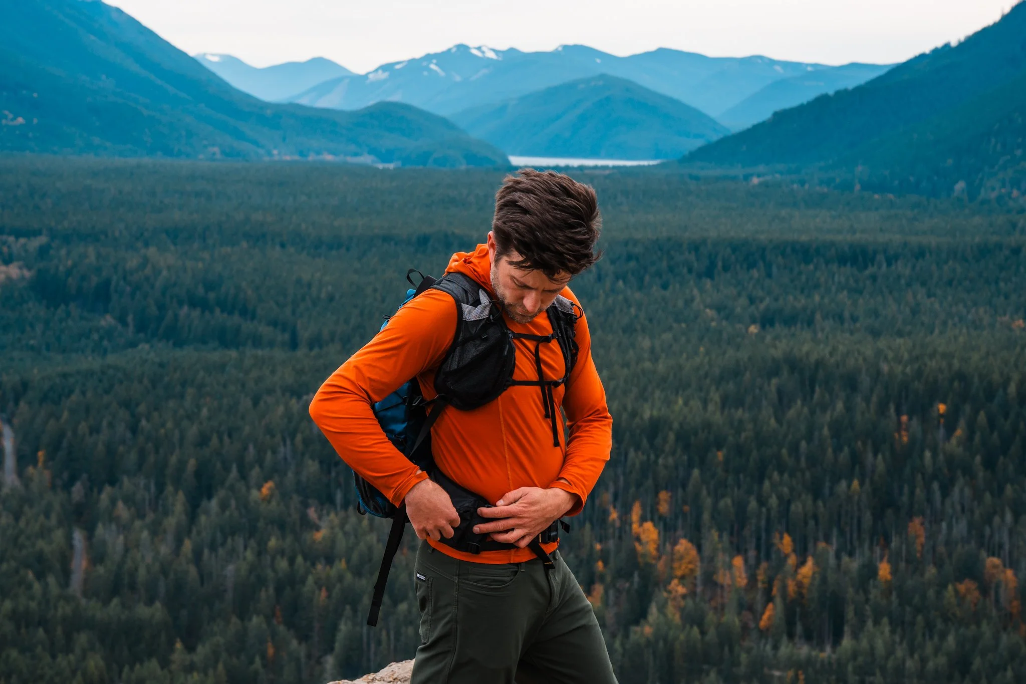 a man standing at a view point on a hike, wearing an orange sun hoodie and backpack with mountains in the background