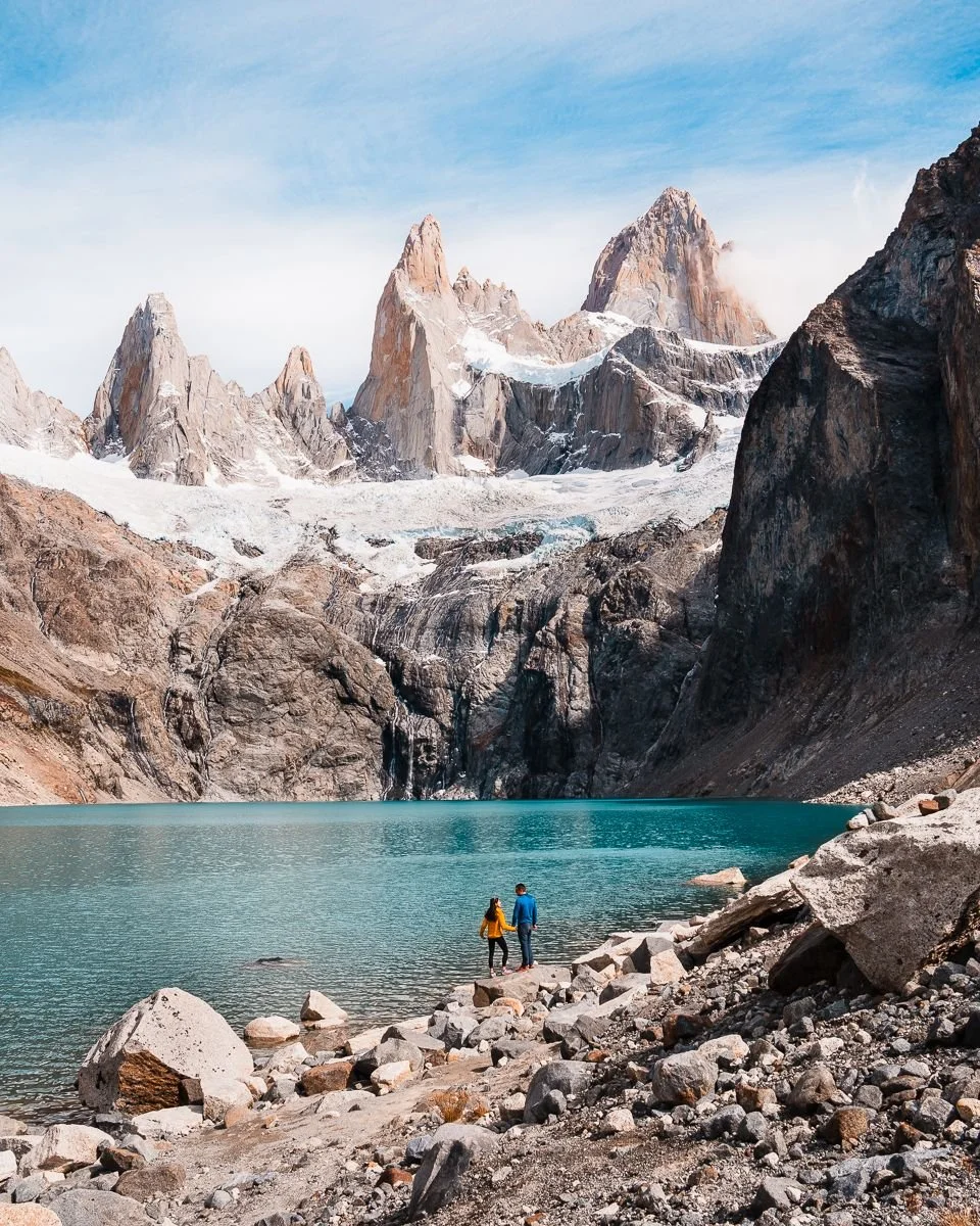 Two people standing on rocky shoreline by a turquoise mountain lake, with snow-capped peaks and rocky cliffs in the background under a blue sky.