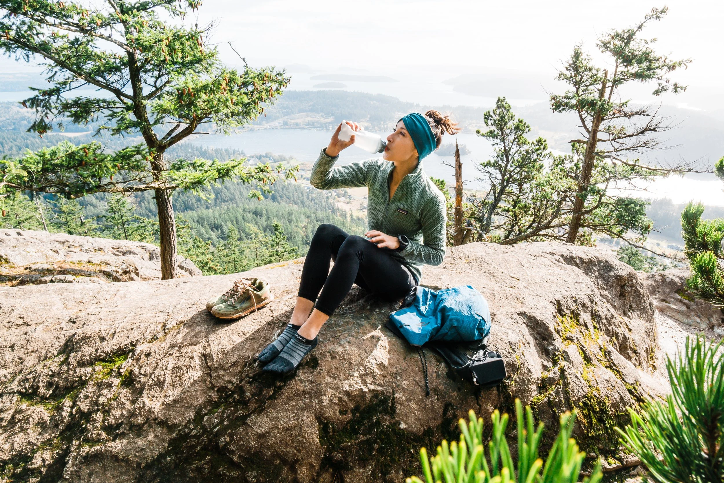 Woman sitting on a large rock in the forest, drinking water from a bottle, with a backpack and shoes nearby, overlooking a scenic view of trees, water, and distant land.