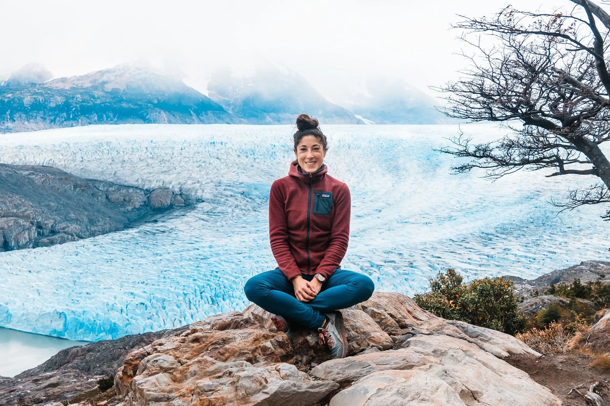 a woman sitting on a rock, wearing a maroon and navy full zip fleece, sitting in front of a massive glacier and icefield in patagonia