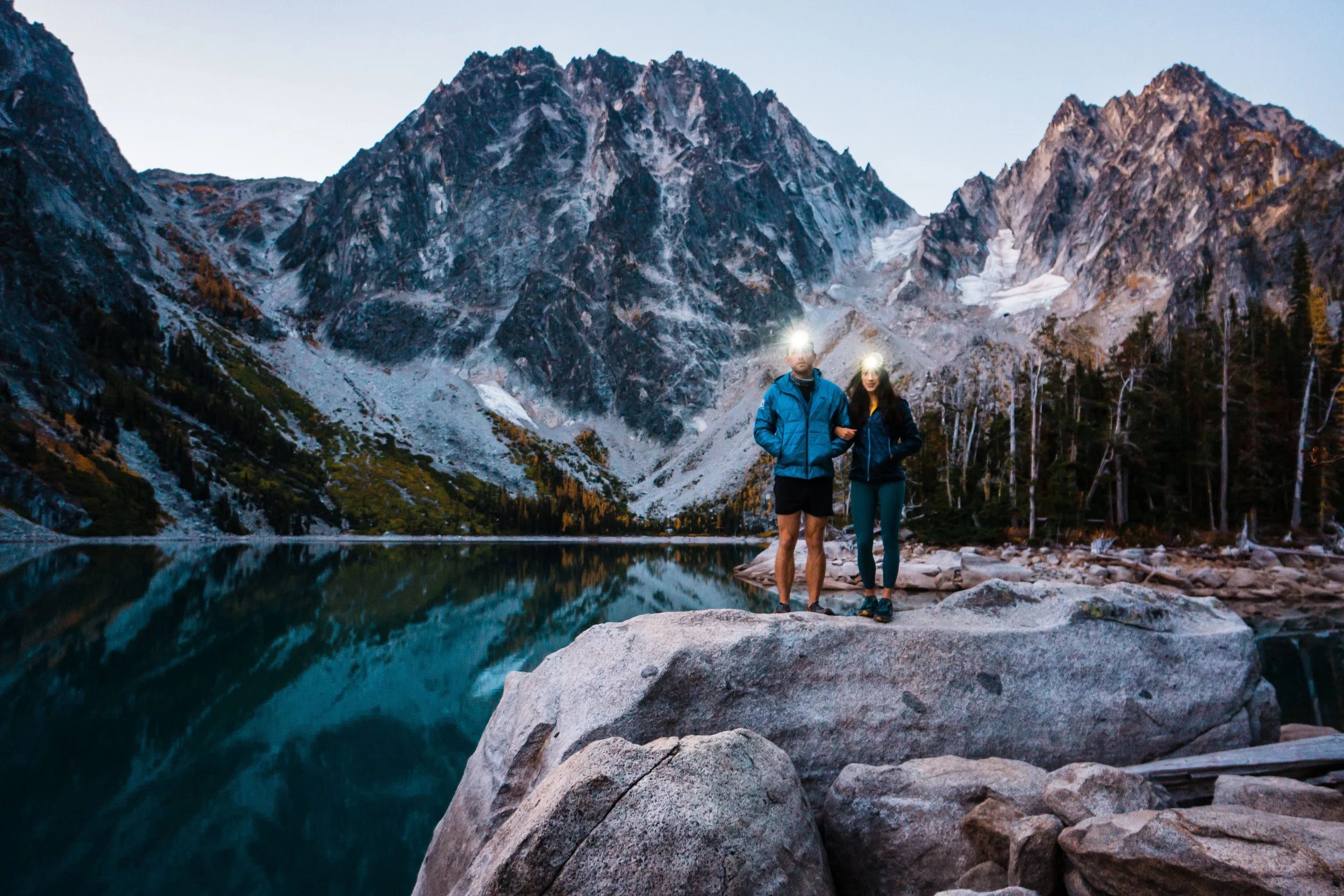 A couple standing on a rock with headlamps on, in front of colchuck lake, a reflective blue lake with a big rocky, dragontail peak behind
