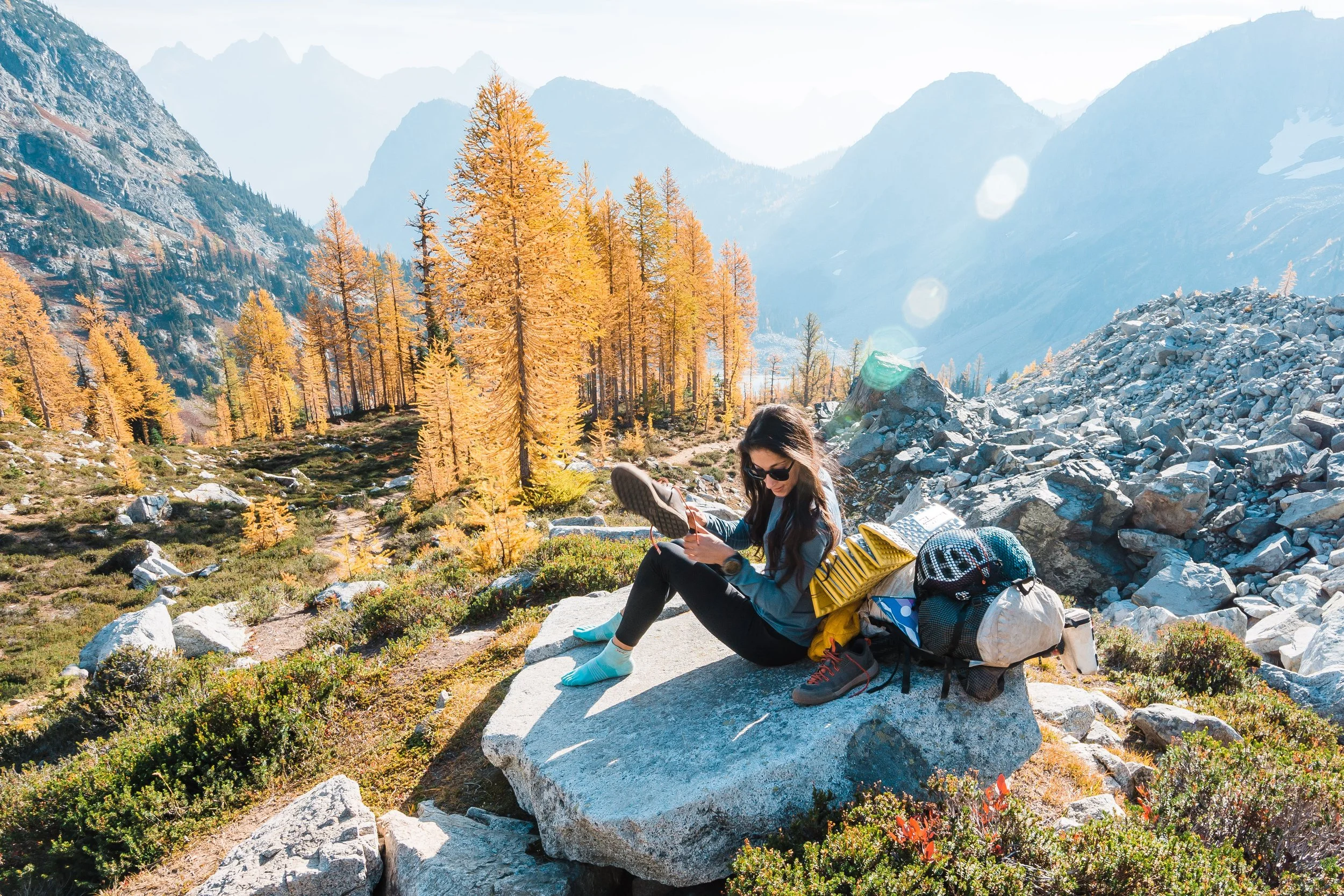 A woman sitting on a large rock in a mountainous landscape with colorful trees, using her phone during daylight.