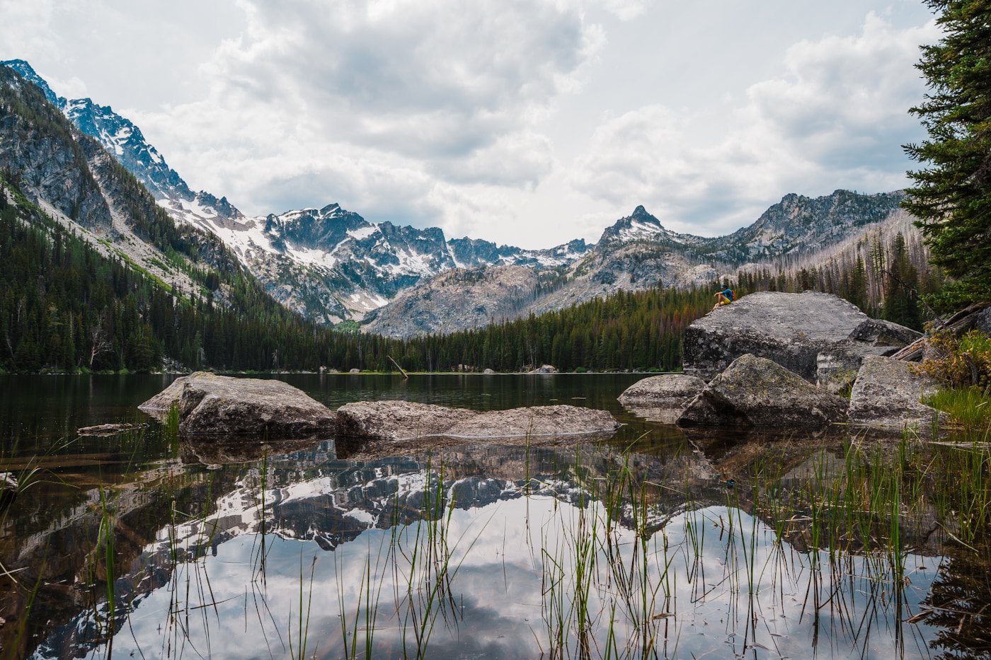views of Stuart Lake in the Enchantments, with mountains reflecting in the lake and a hiker sitting on a big boulder on the lake shore