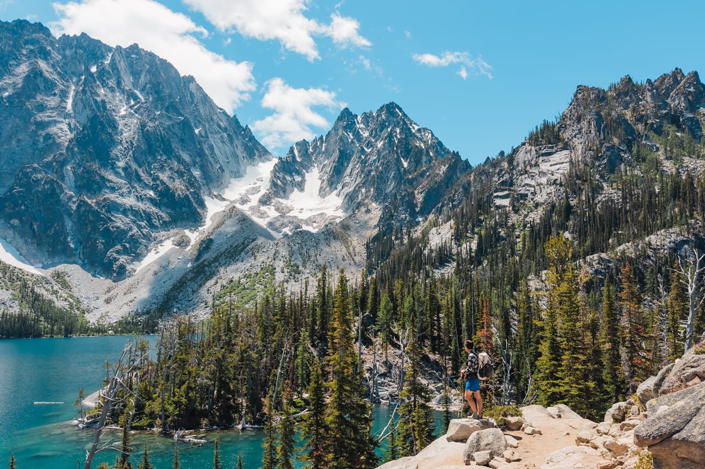 a tiny backpacker standing on a rock looking out at colchuck lake with colchuck peak in the background