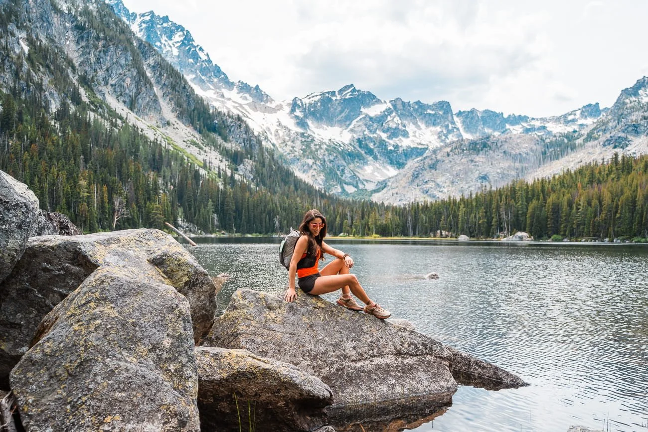 a woman sitting on a rock at a lake shore with water and mountains in the background