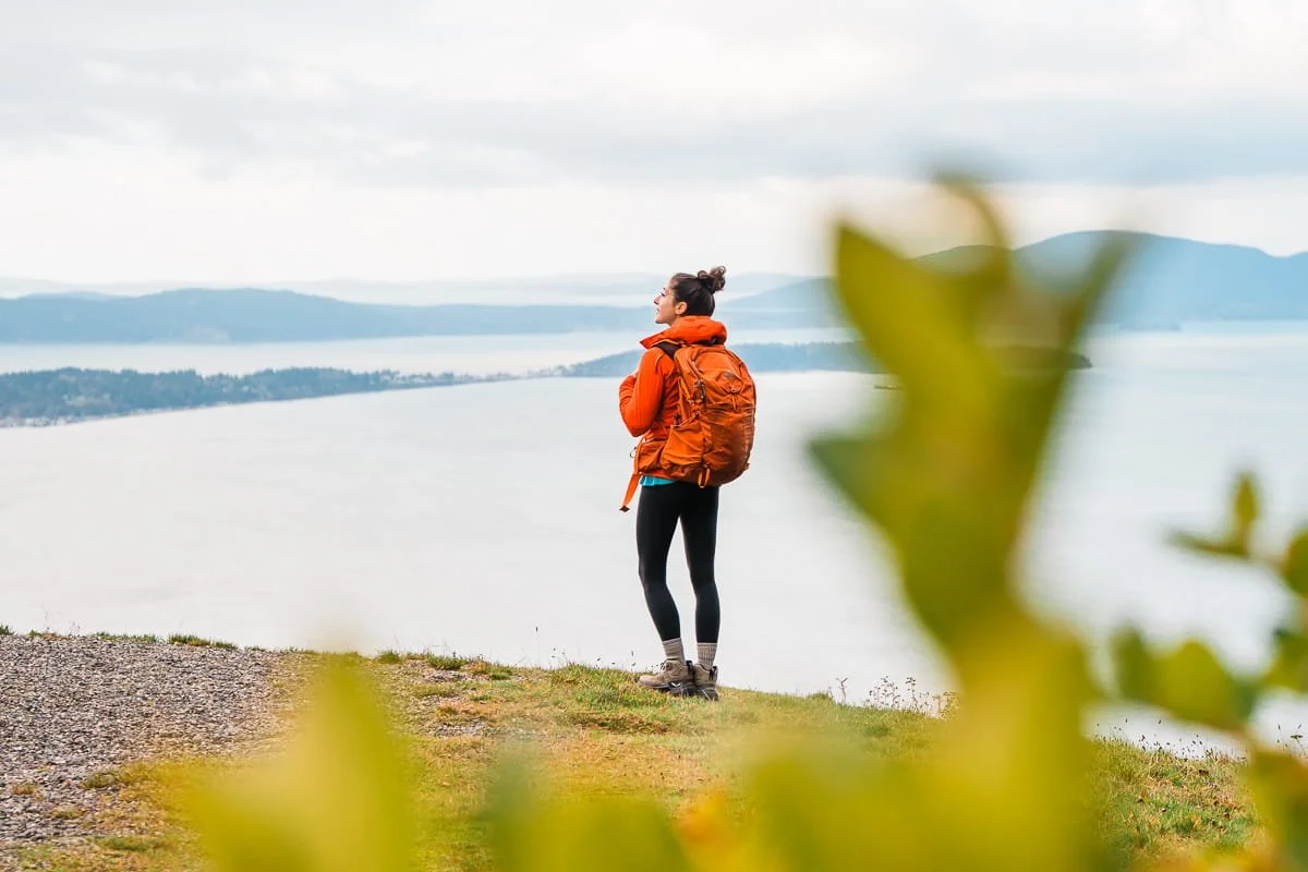 A woman with an orange day pack standing and looking out at a view of the sound in washington