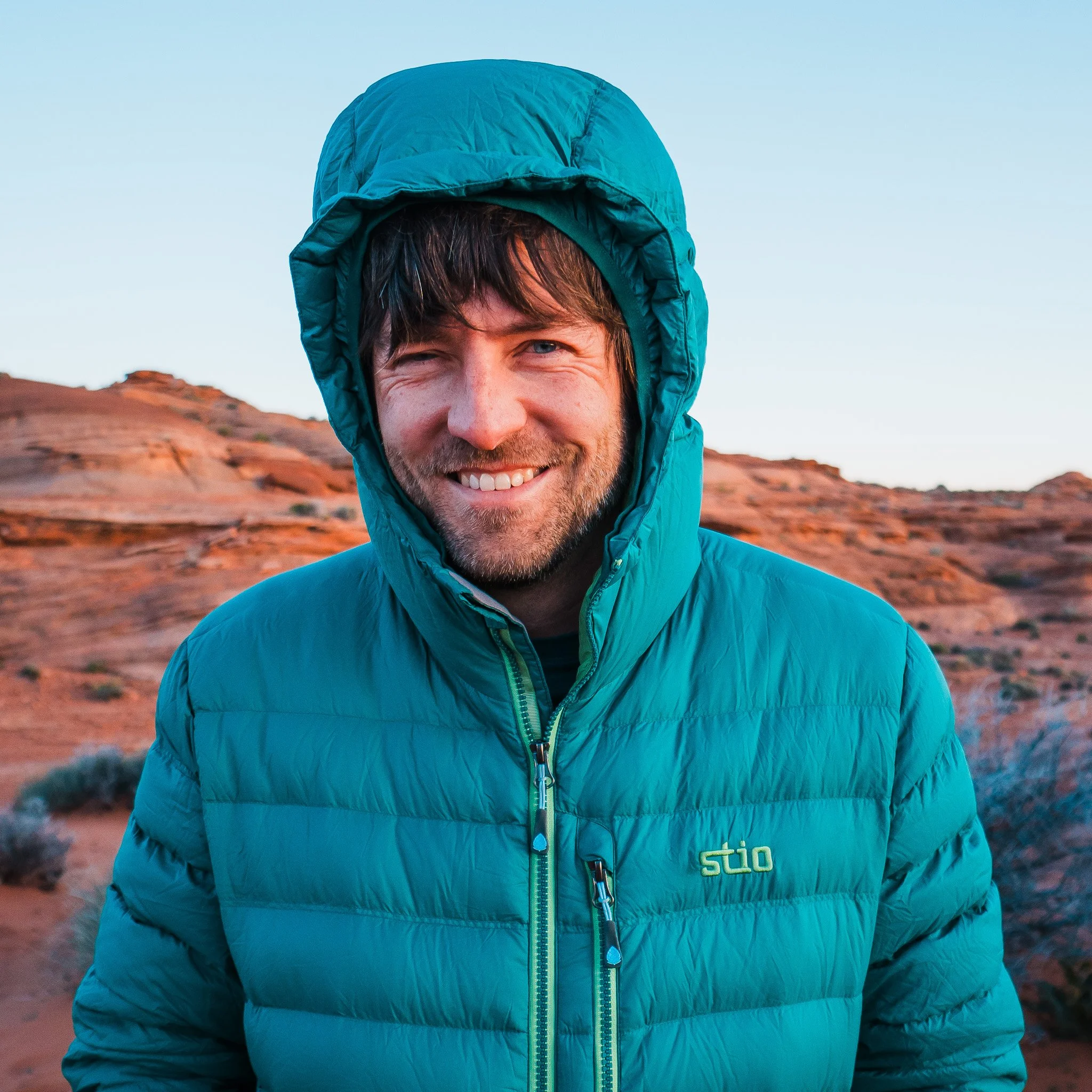 A man wearing a greenish blue down jacket with the hood up, standing in the desert in winter