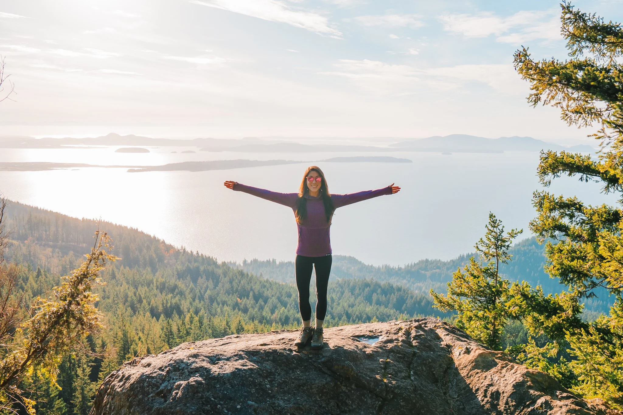 A woman standing on a rock in front of a view of the sound, with arms outstretched, wearing a purple fleece
