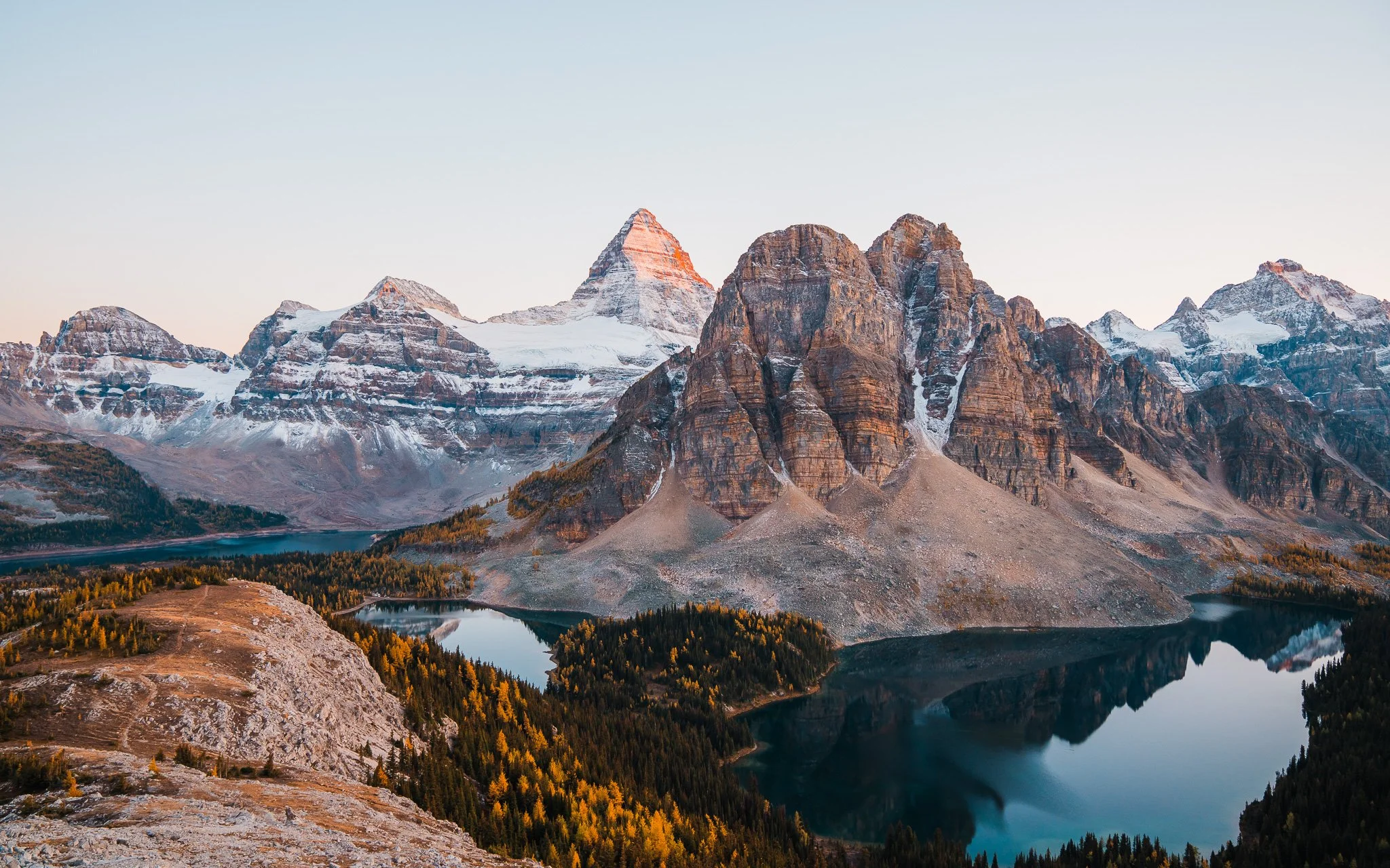 a pointy, snow dusted peak with another rocky peak next to it. Looking down at two lakes and trees beneath.