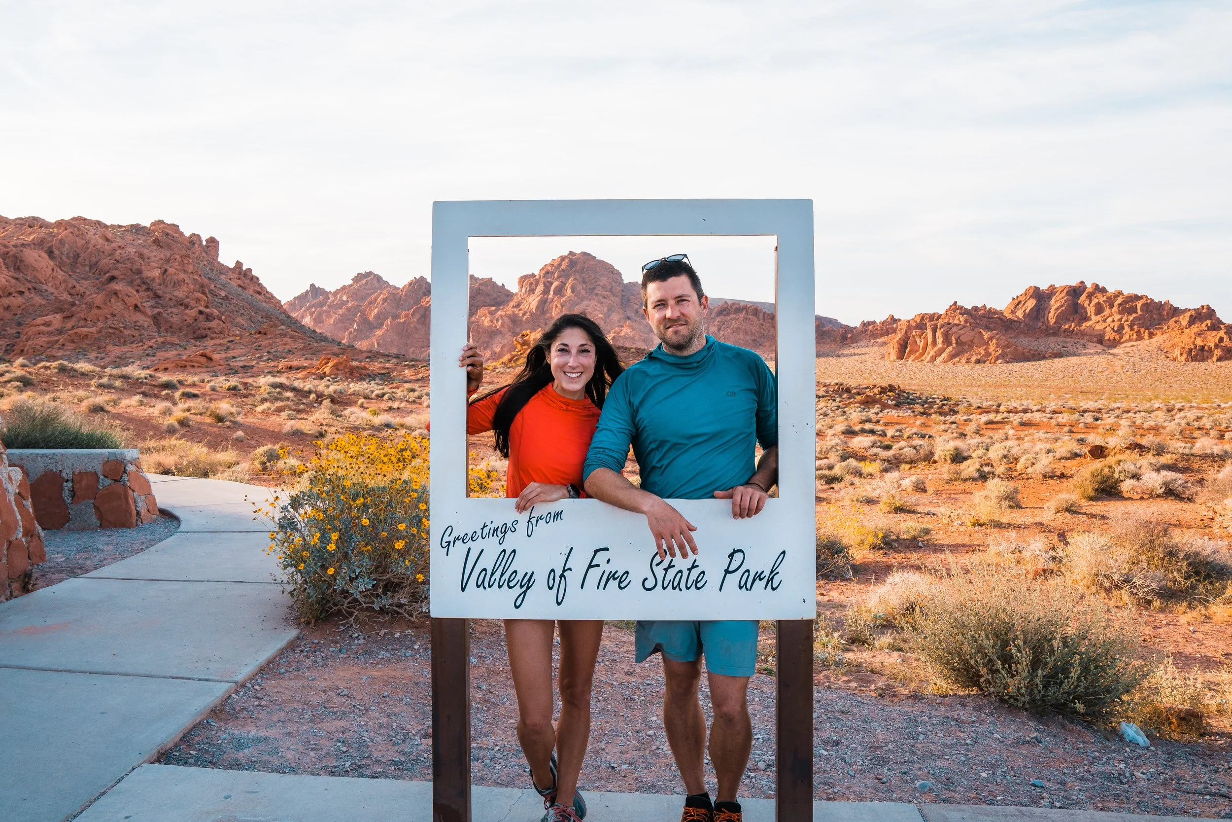 A smiling woman in an orange shirt and a man in a blue shirt and gray shorts stand behind a white frame with the words "Greetings from Valley of Fire State Park," posing for a photo in a desert landscape with rocky red mountains and sparse vegetation