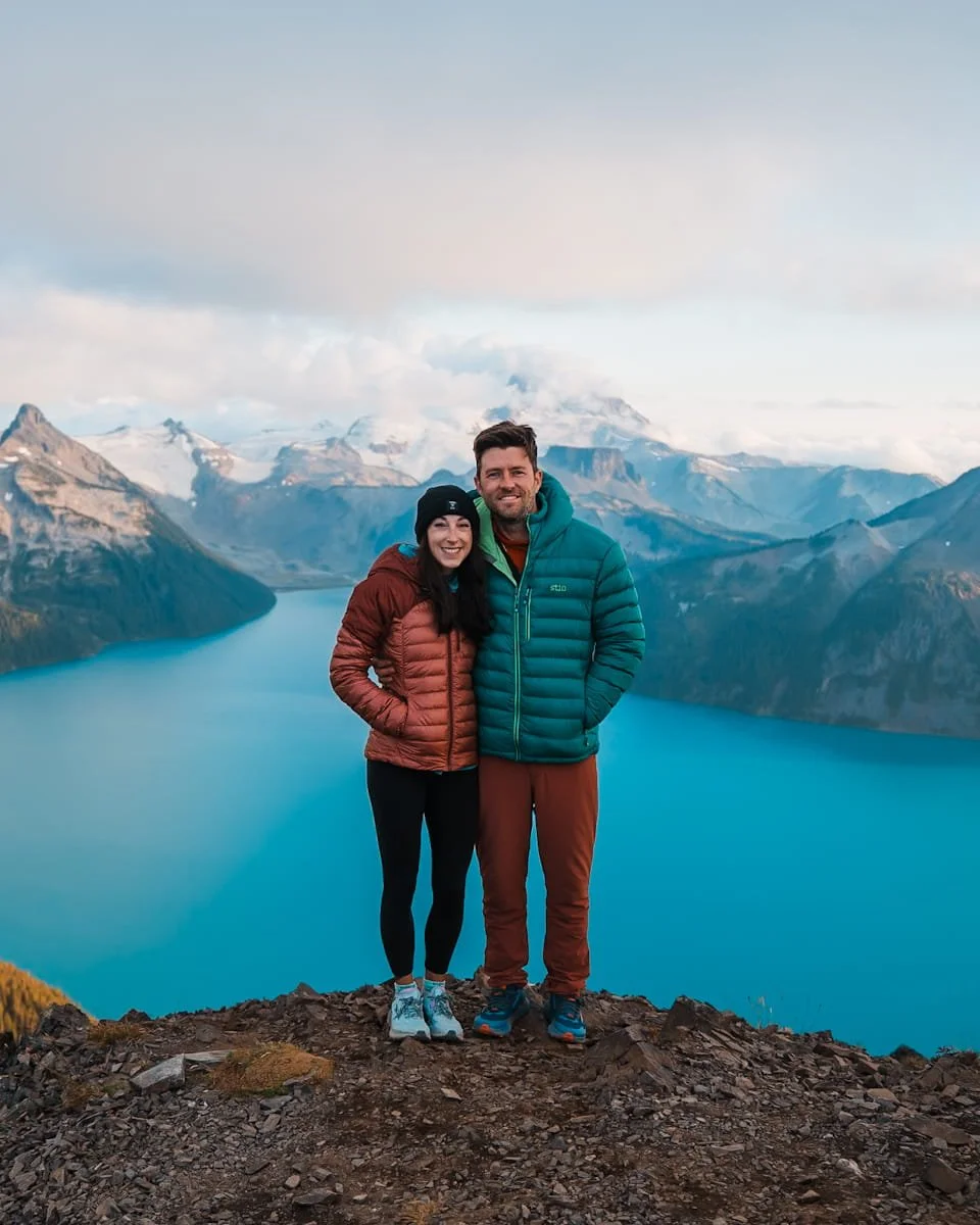 A couple smiling at the camera, in down jackets, at a hiking view point with glaciated mountains and a bright blue lake in the background