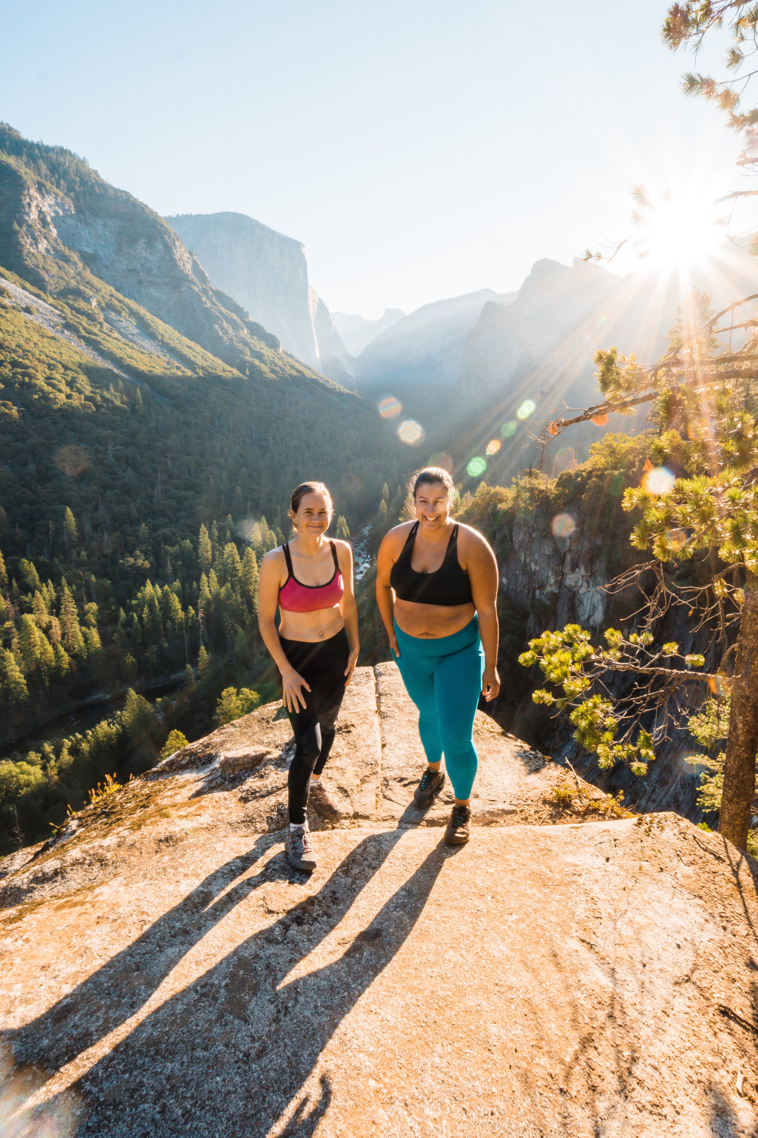 Two women in athletic clothing hiking on a mountain trail during sunset, with a scenic view of mountains and trees in the background.